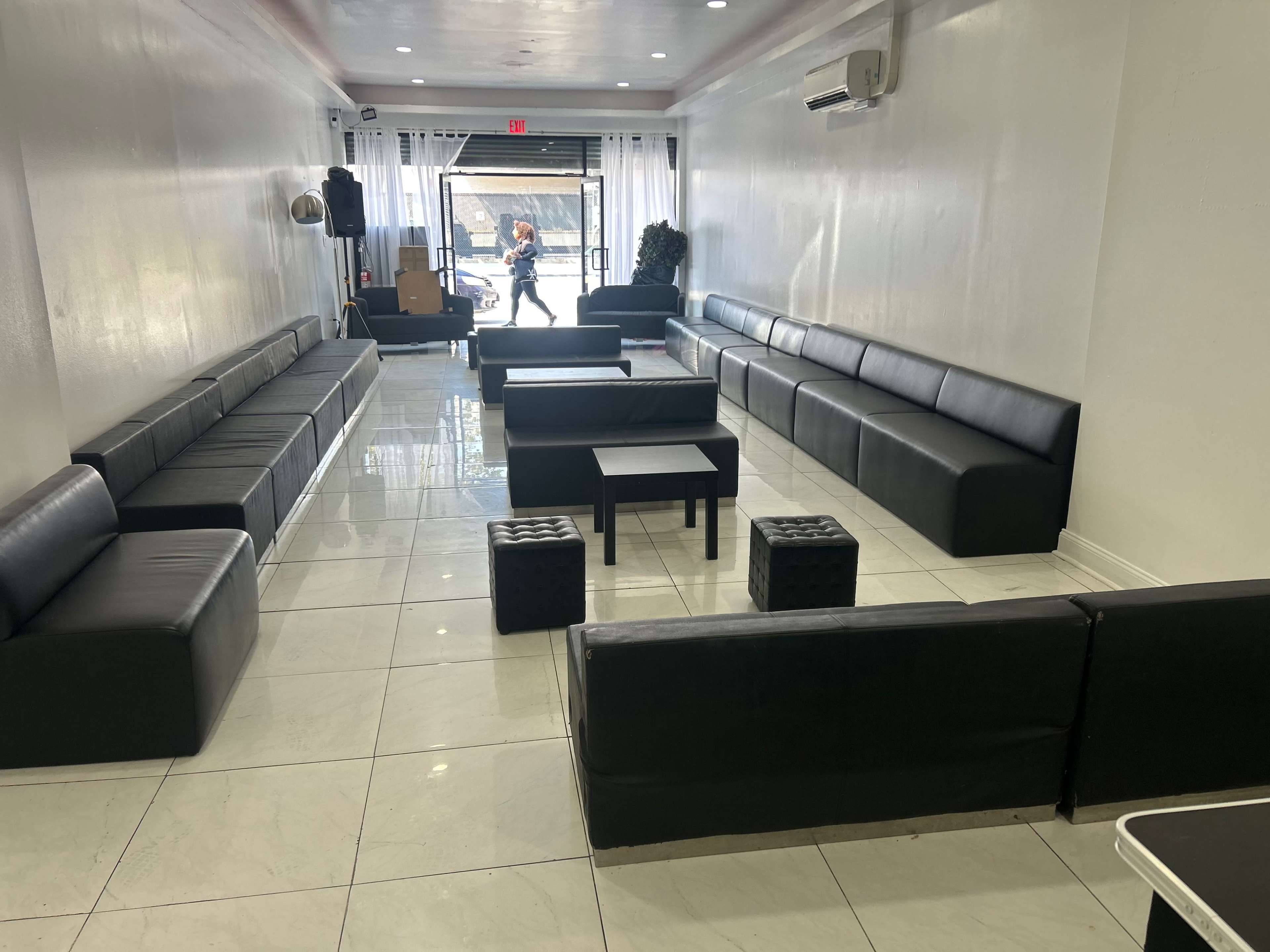 The image shows a minimalist indoor space furnished with black couches and small tables, featuring a large window with natural light.
