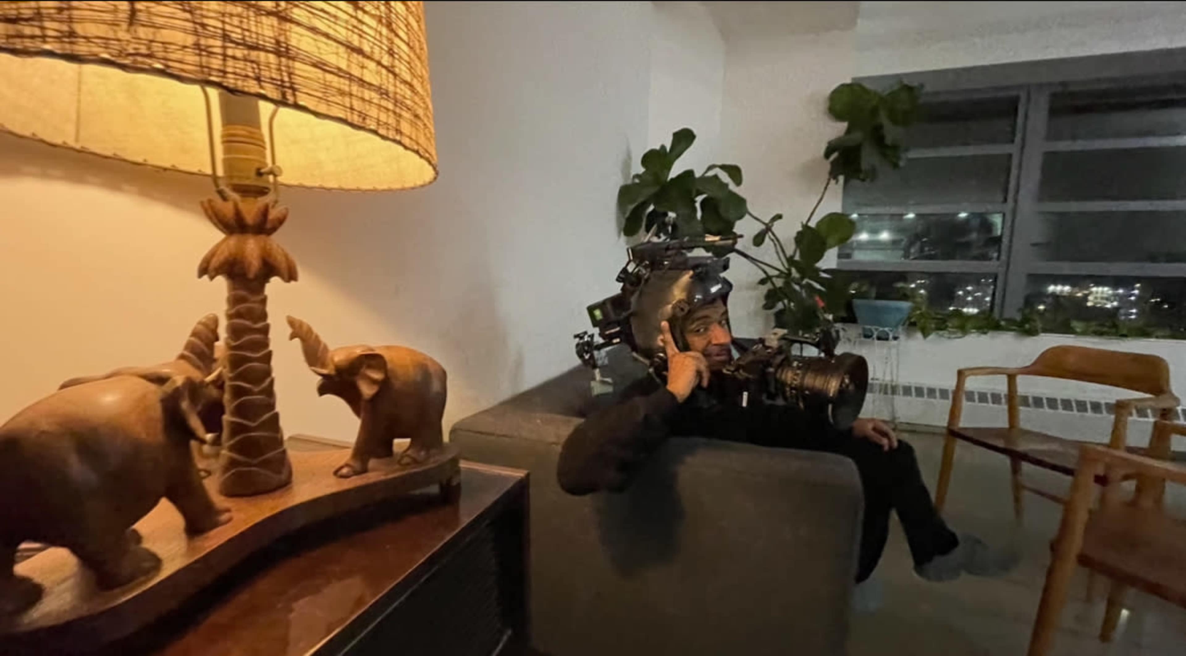 A person wearing a camera rig sits on a couch next to a lamp and decorative elephant figurines in a well-lit room.