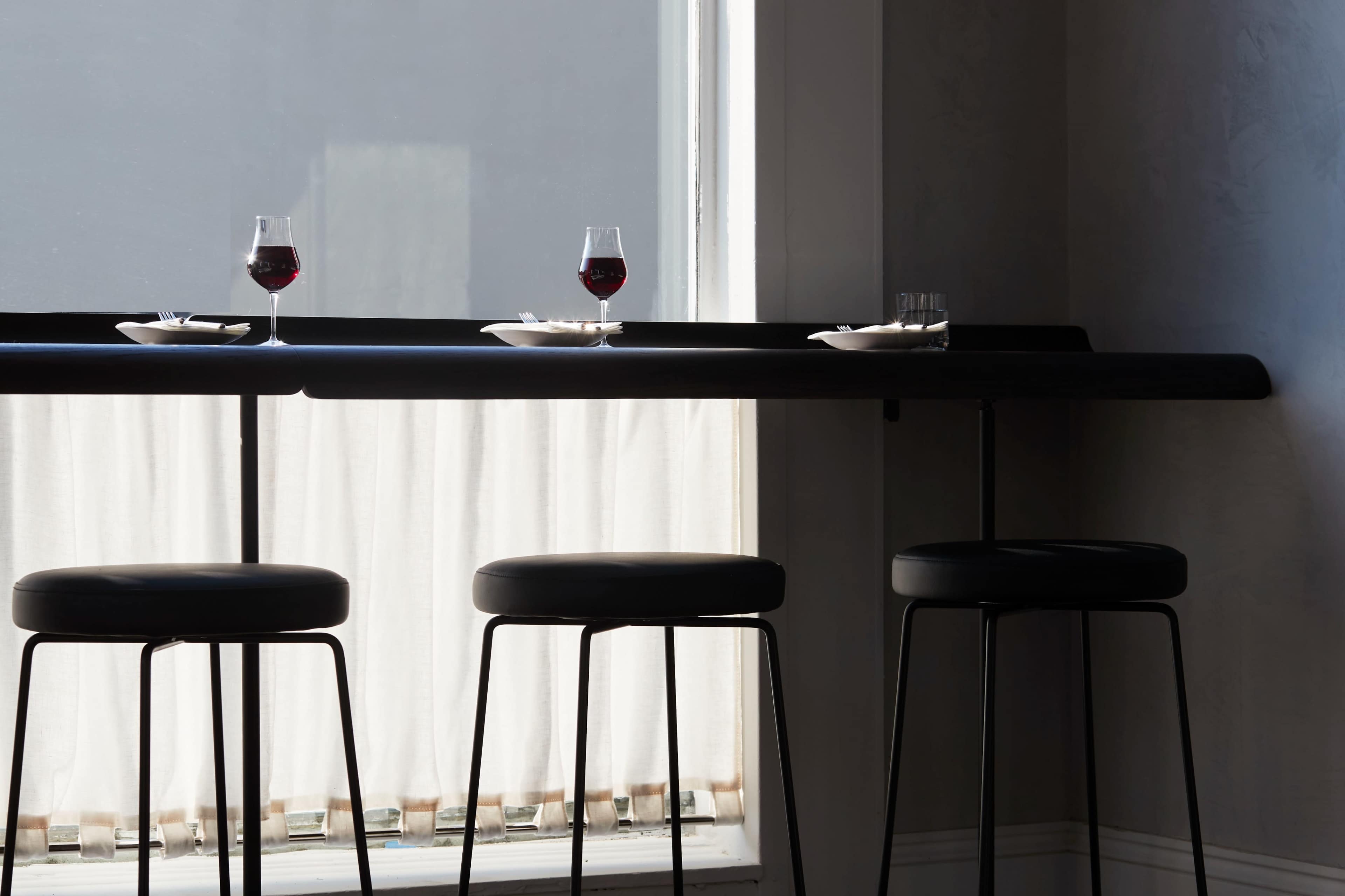 A dimly lit bar area features three black stools with glasses of red wine and small plates on a dark table near a window with sheer curtains.
