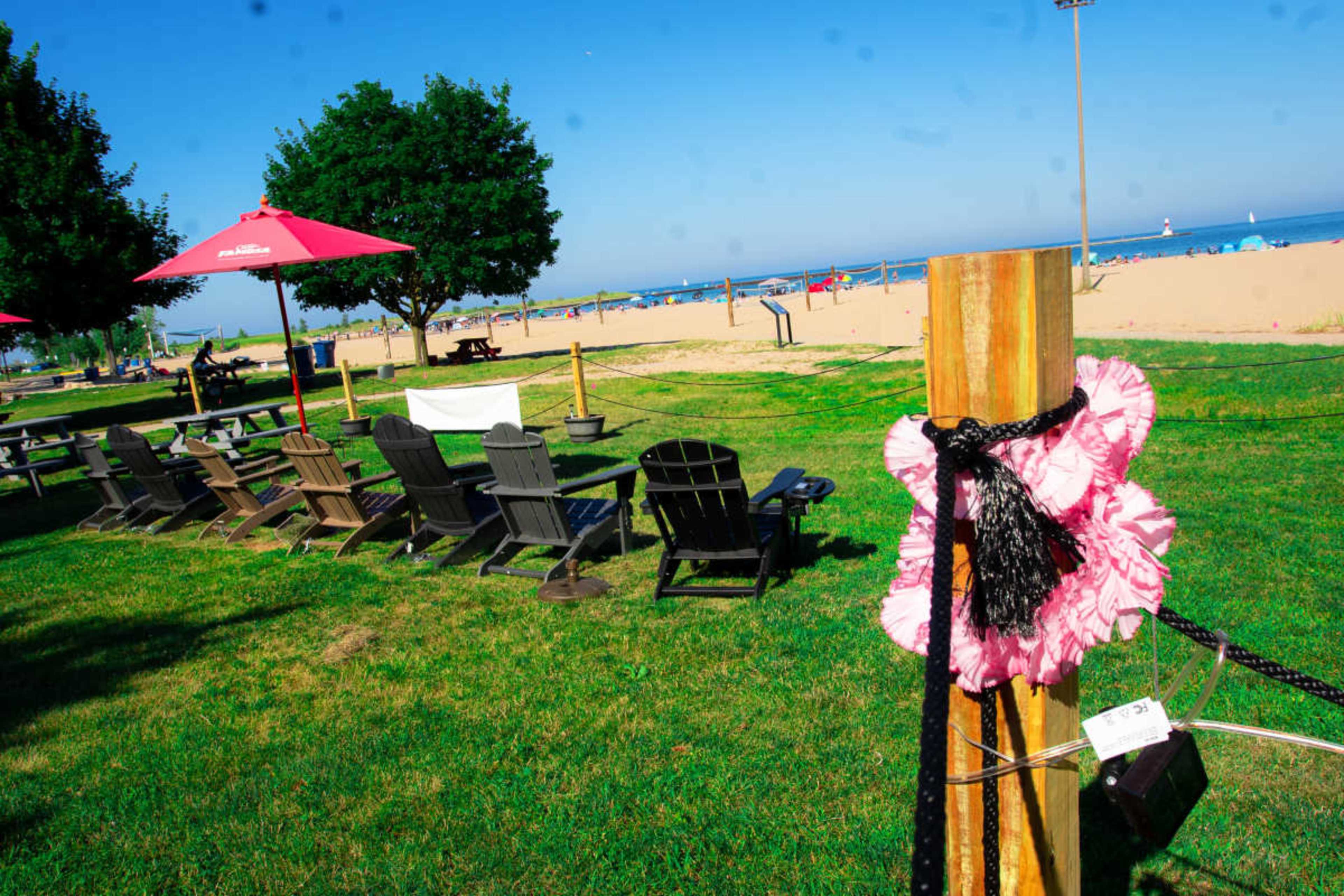The scene depicts a sunny beachside park with rows of wooden chairs under red umbrellas and a sandy beach in the background.