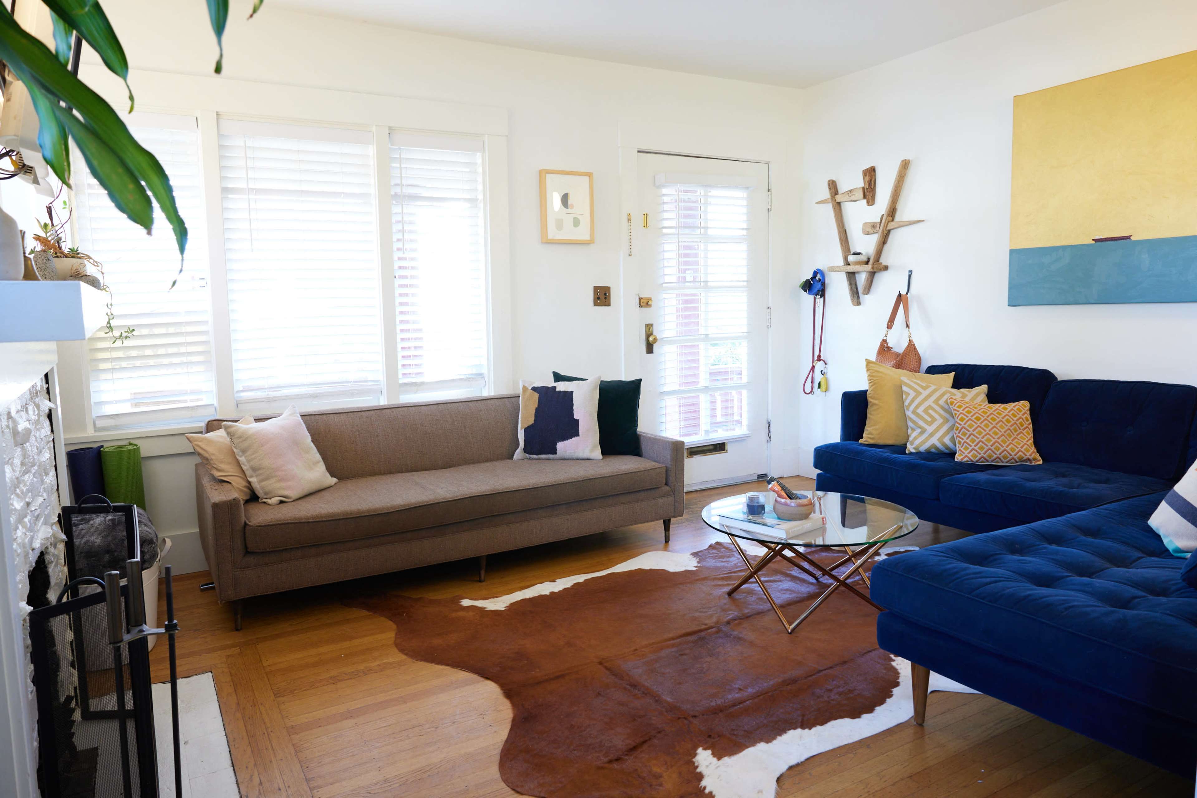A living room features a brown sofa and a blue sectional couch, a glass coffee table on a cowhide rug, and a door leading outside.