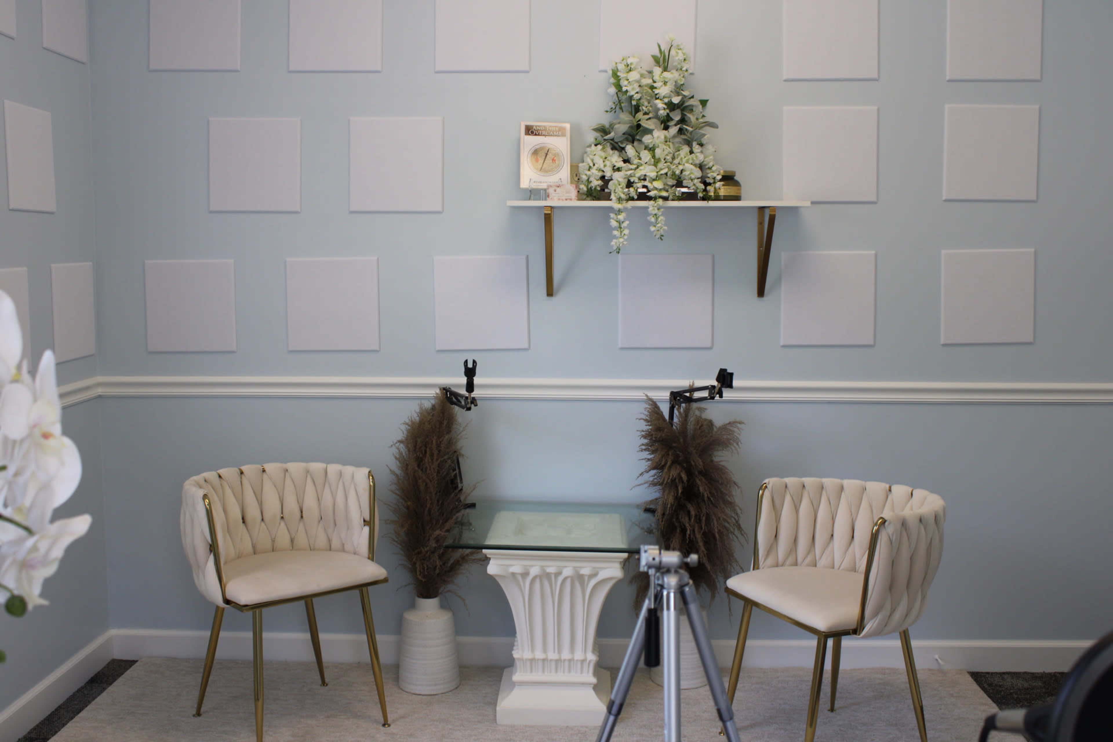 The image shows a well-arranged room with light blue walls, two white upholstered chairs with gold legs, a glass table, and decorative elements on a shelf, including plants and a round object.