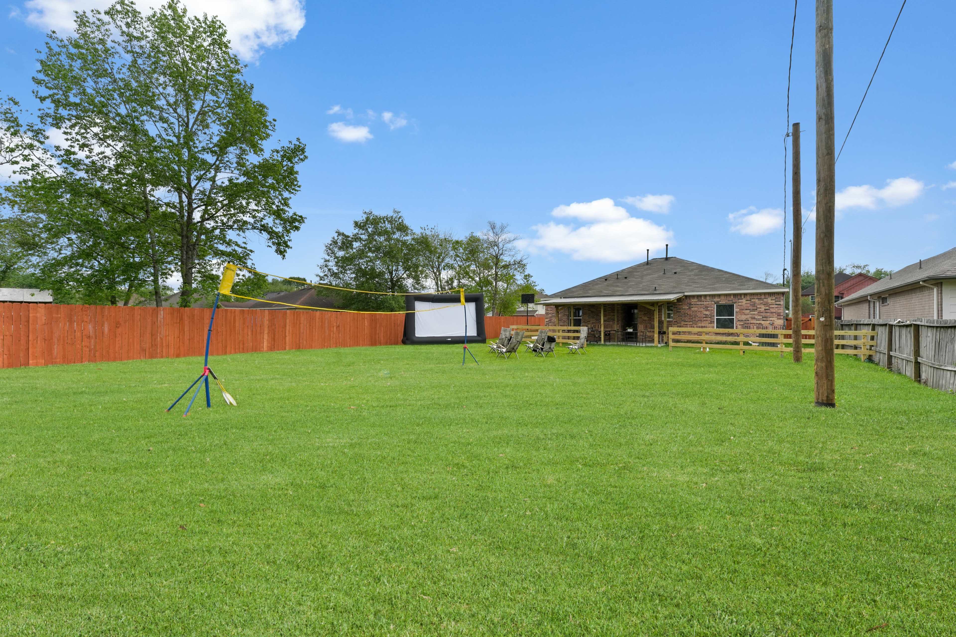 The image shows a spacious backyard with a volleyball net, a movie screen setup, and a seating area, surrounded by wooden fencing.