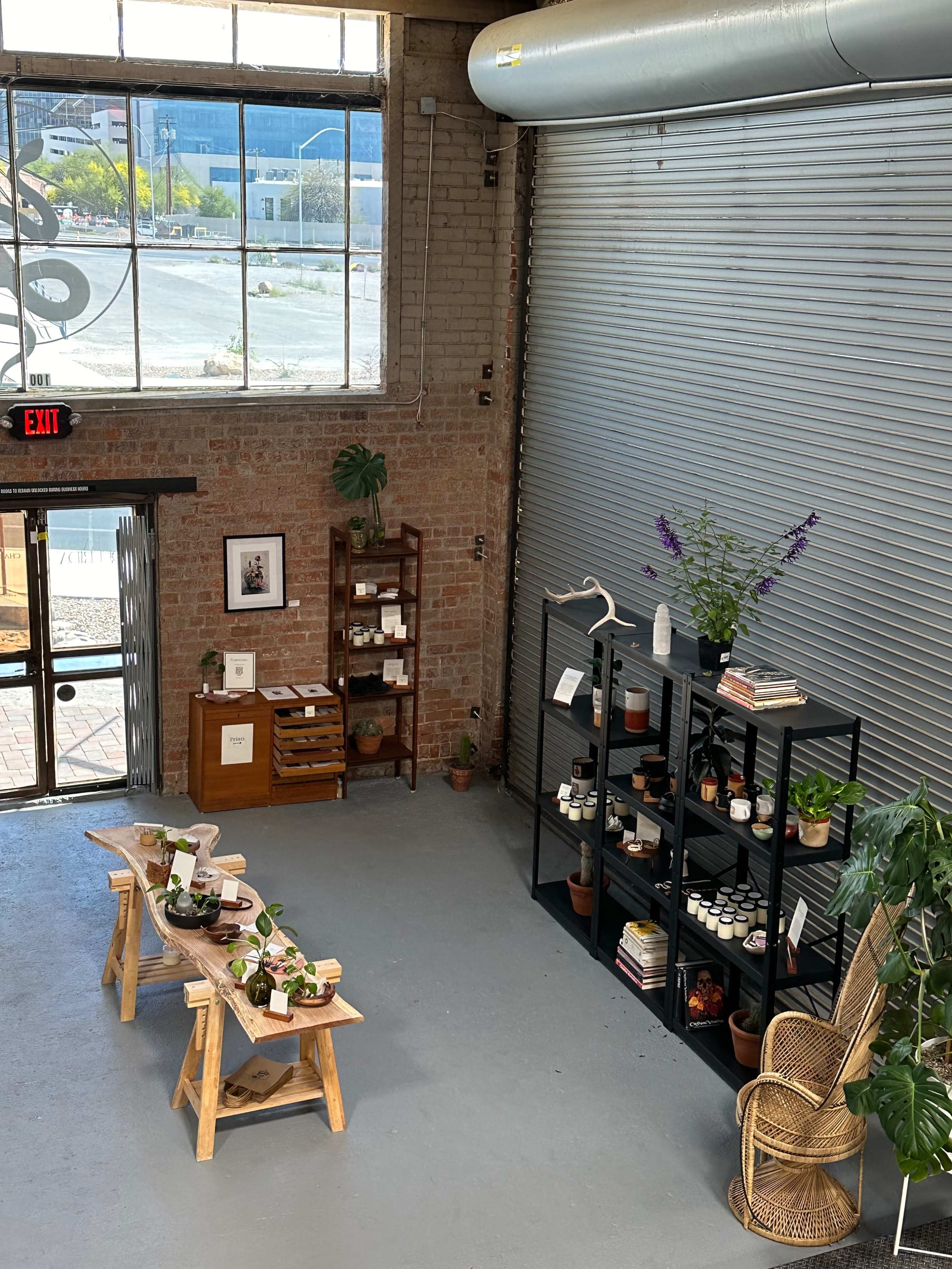 A spacious interior featuring wooden tables with plants, shelves filled with jars and books, and large windows overlooking an outdoor area.