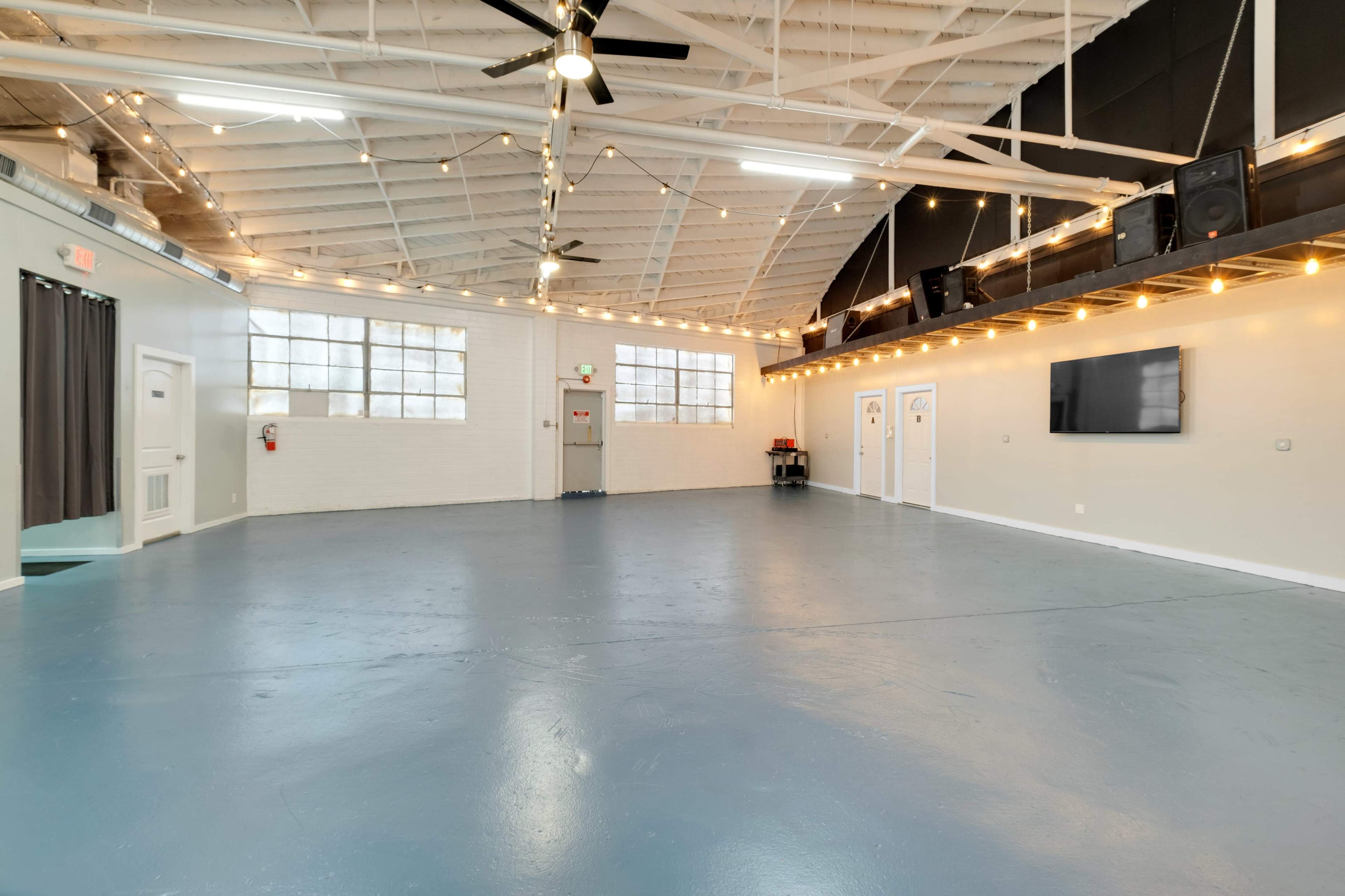 A spacious, well-lit interior with high ceilings, large windows, and a polished floor, featuring a TV mounted on the wall and doors lining one side.
