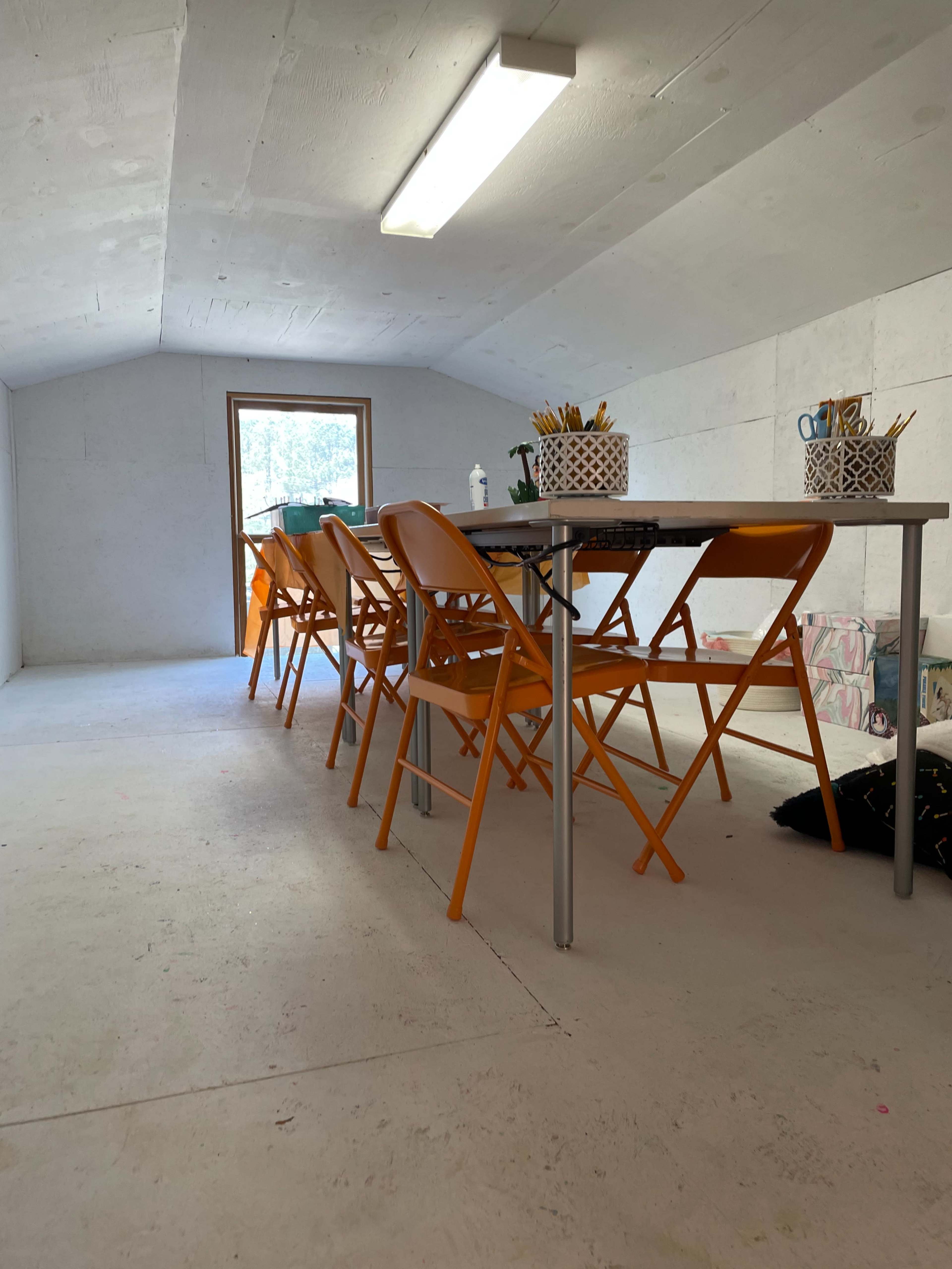A spacious, light-filled attic room features a long table surrounded by orange folding chairs.