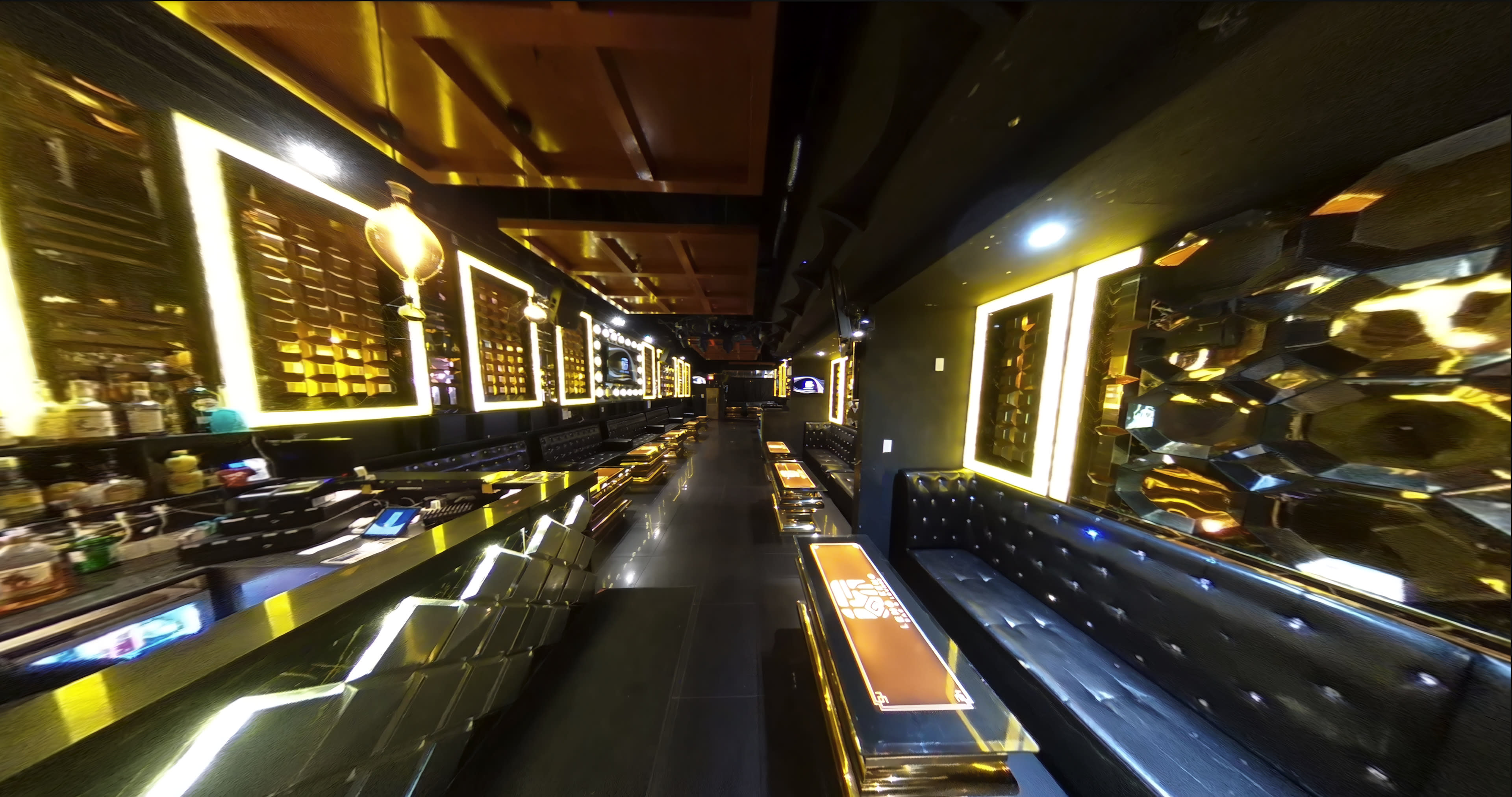 A modern bar interior features a long, sleek counter with illuminated accents and seating along the walls, framed by mirrored panels and warm lighting.