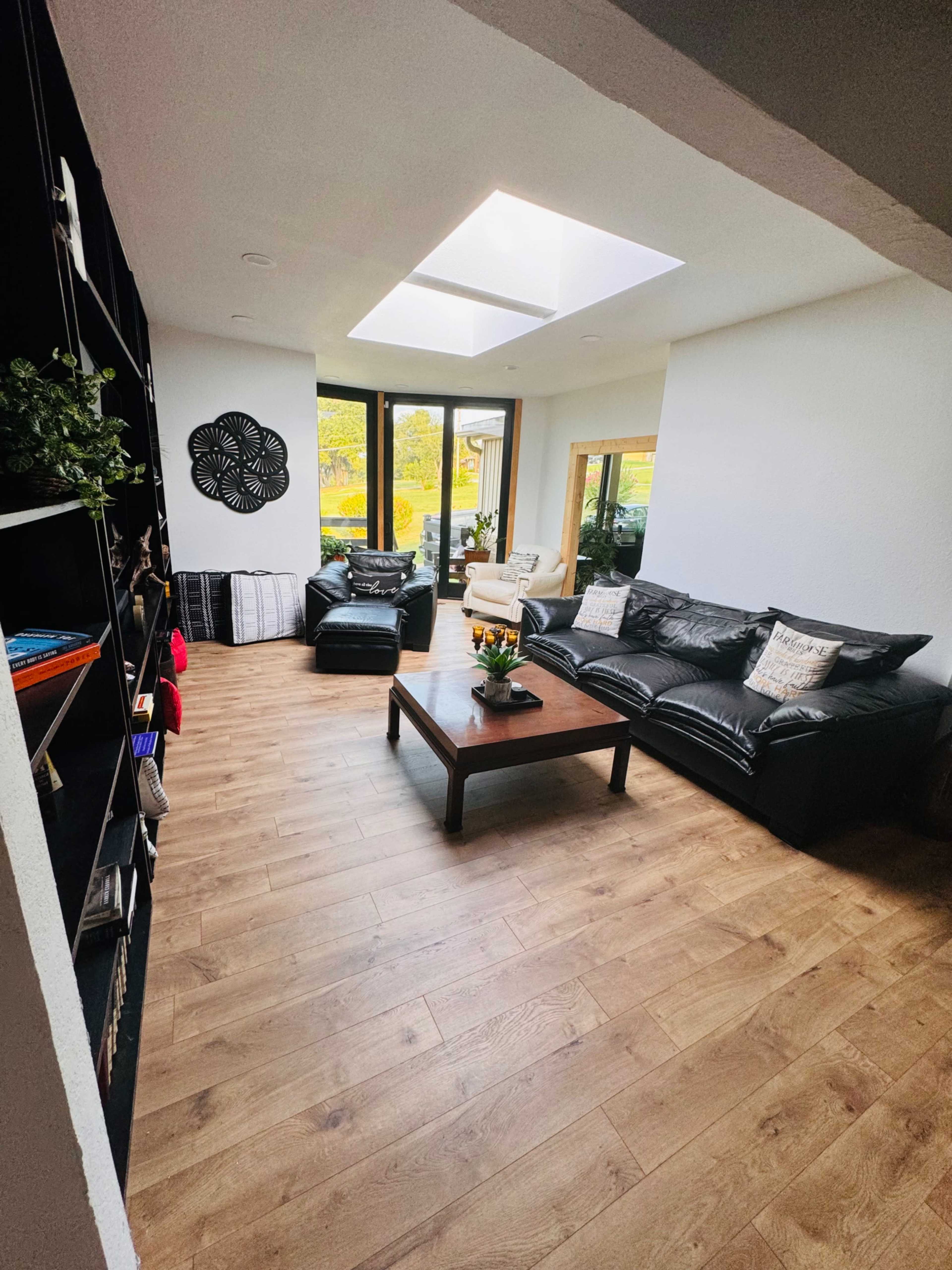 The living room features black leather sofas, a wooden coffee table, and large windows allowing natural light to enter the space.