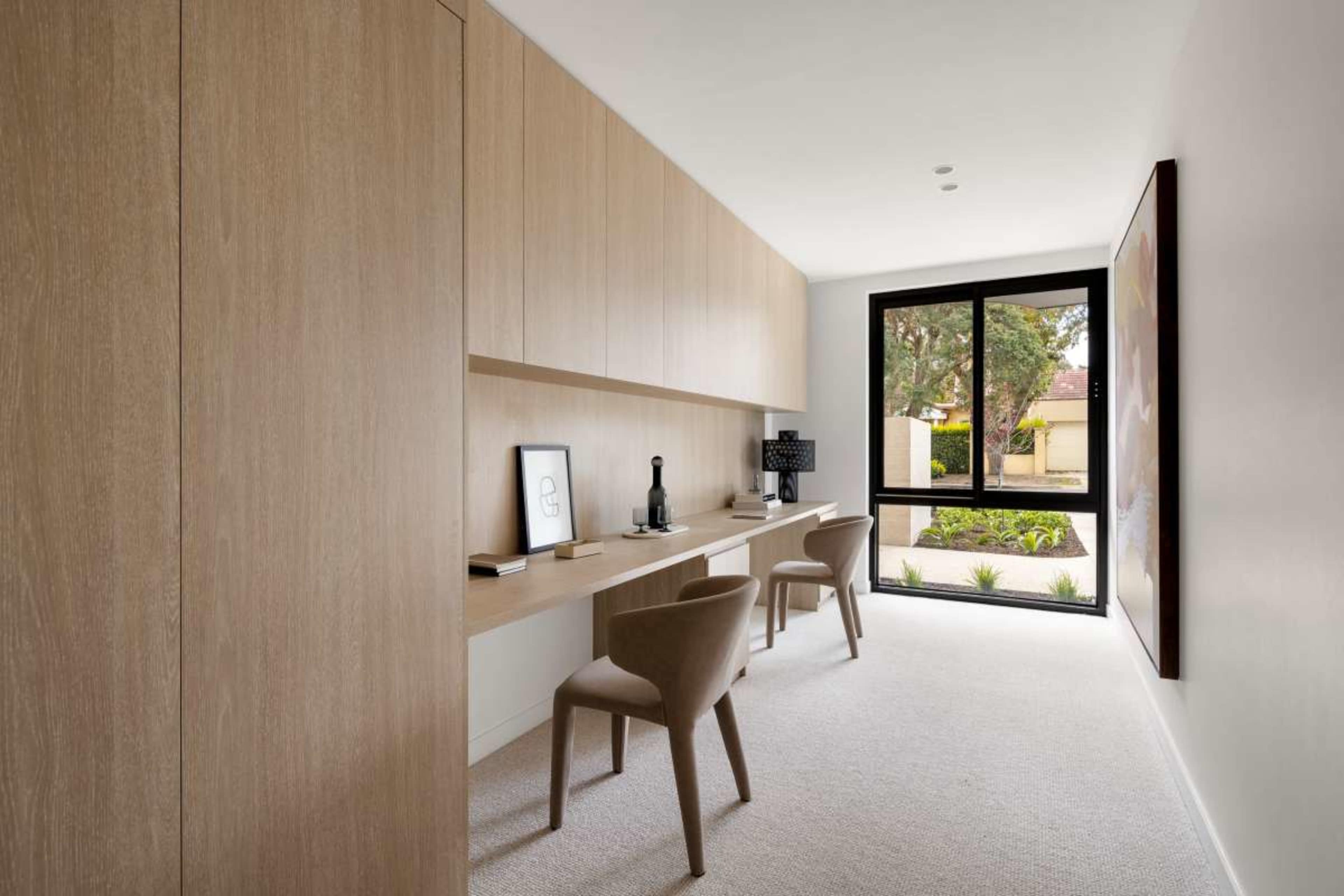The image shows a modern home office with wooden cabinetry, a long desk with a lamp and a computer setup, and two chairs facing large windows that let in natural light.