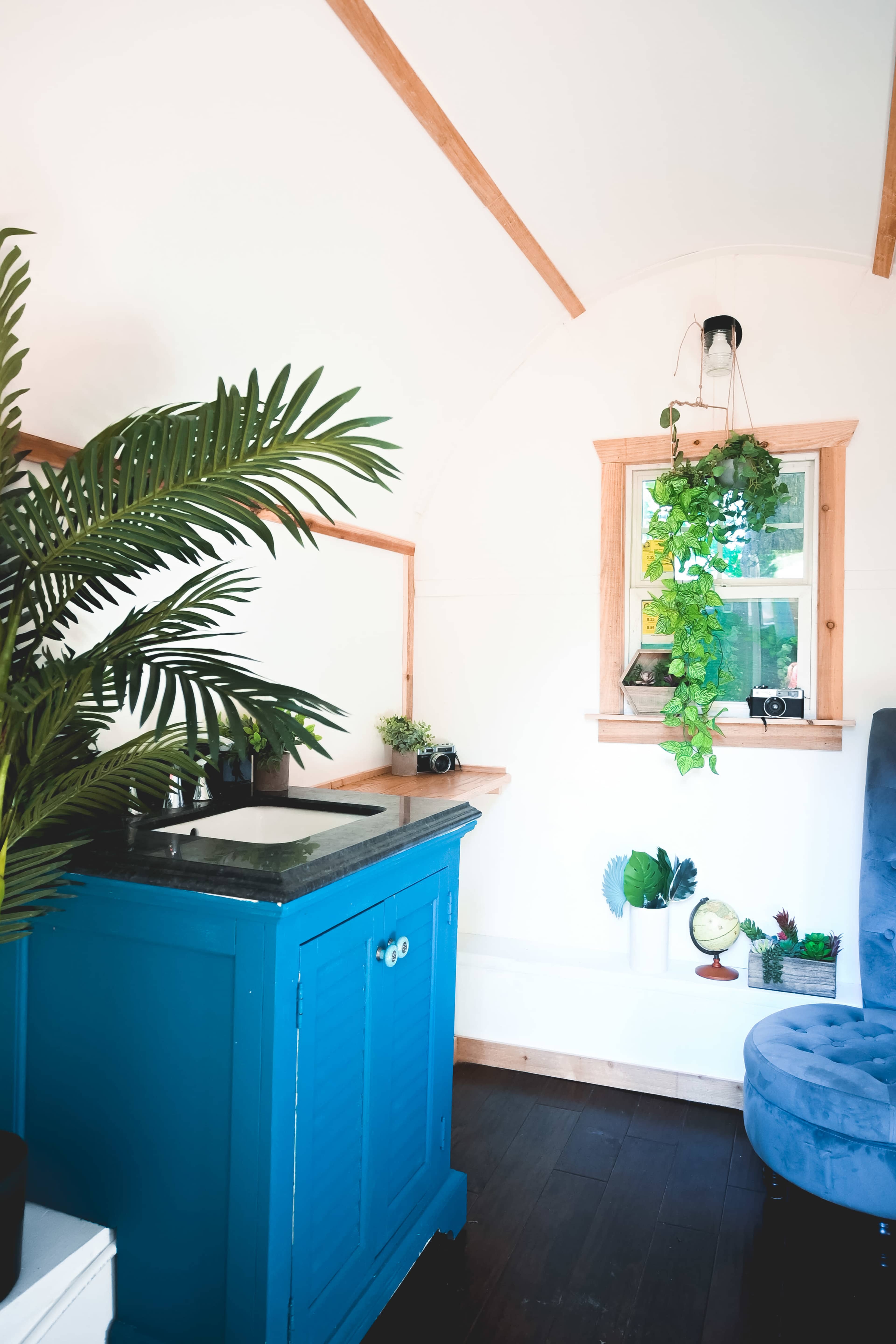 The interior of a small living space featuring a blue kitchen cabinet, a window with hanging plants, and a blue chair.