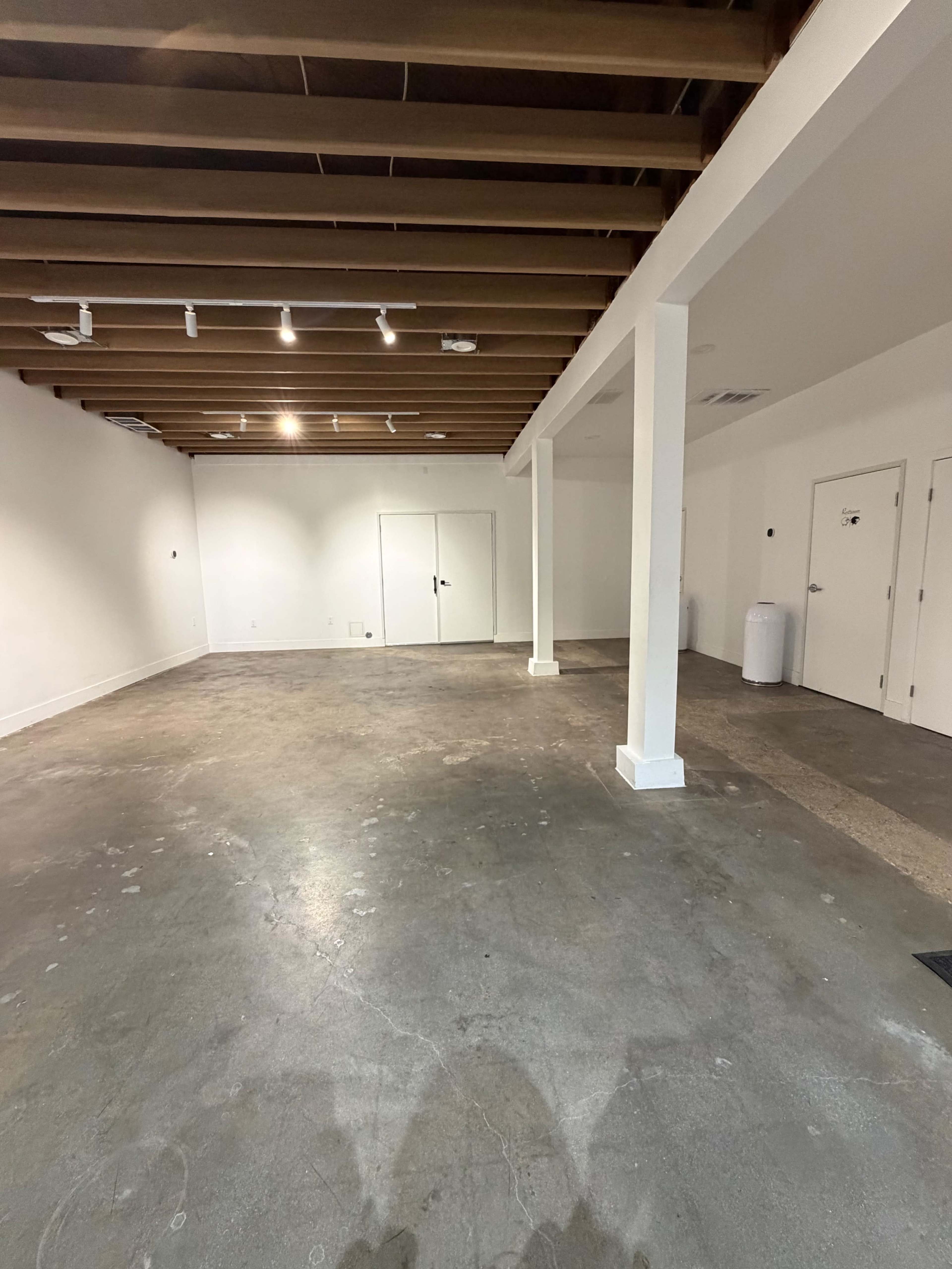 The image shows a spacious, empty room with a concrete floor, white walls, and exposed wooden beams on the ceiling.