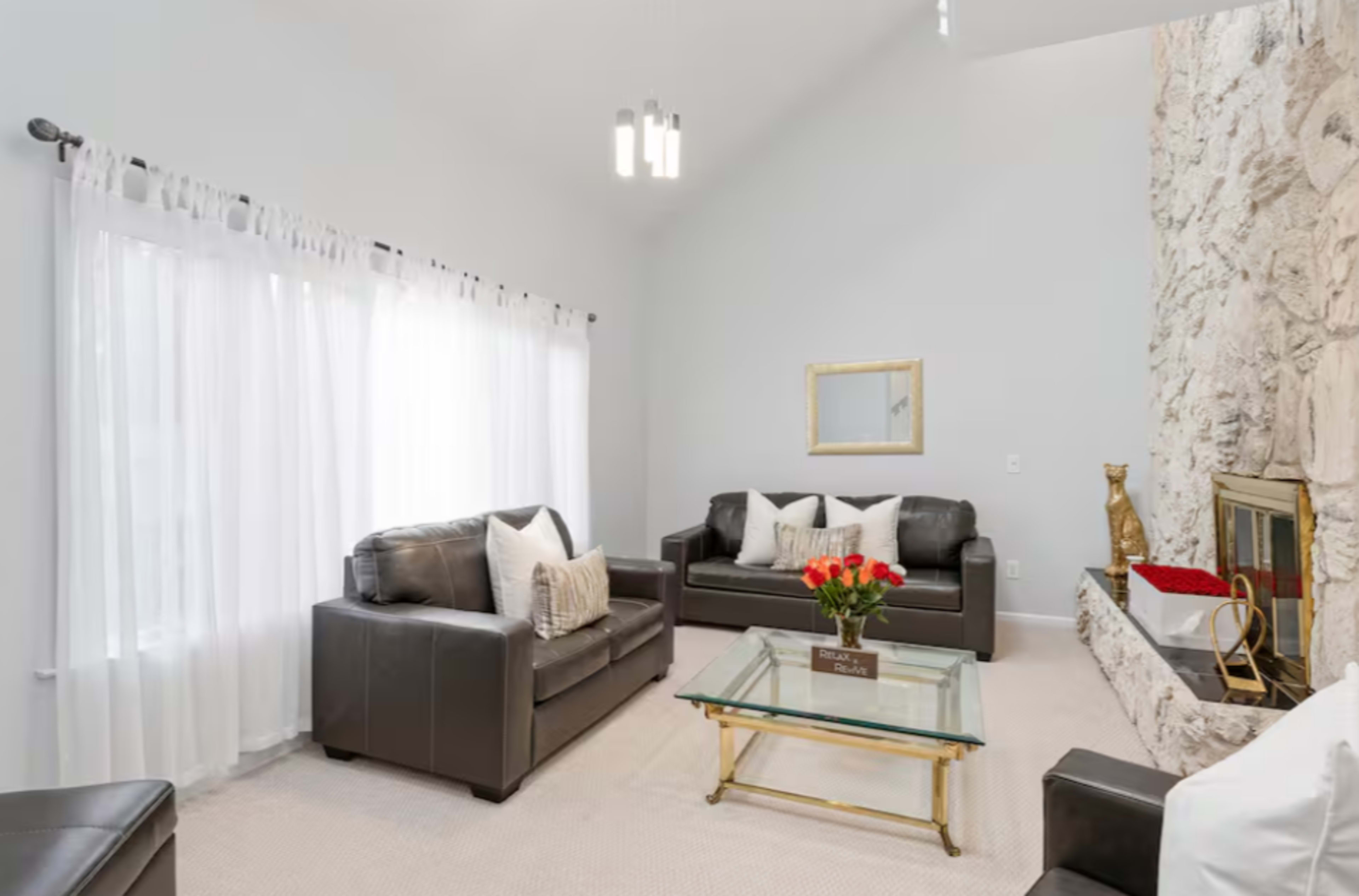 The image shows a modern living room with two dark leather sofas, a glass coffee table, and a stone fireplace, illuminated by natural light through sheer white curtains.
