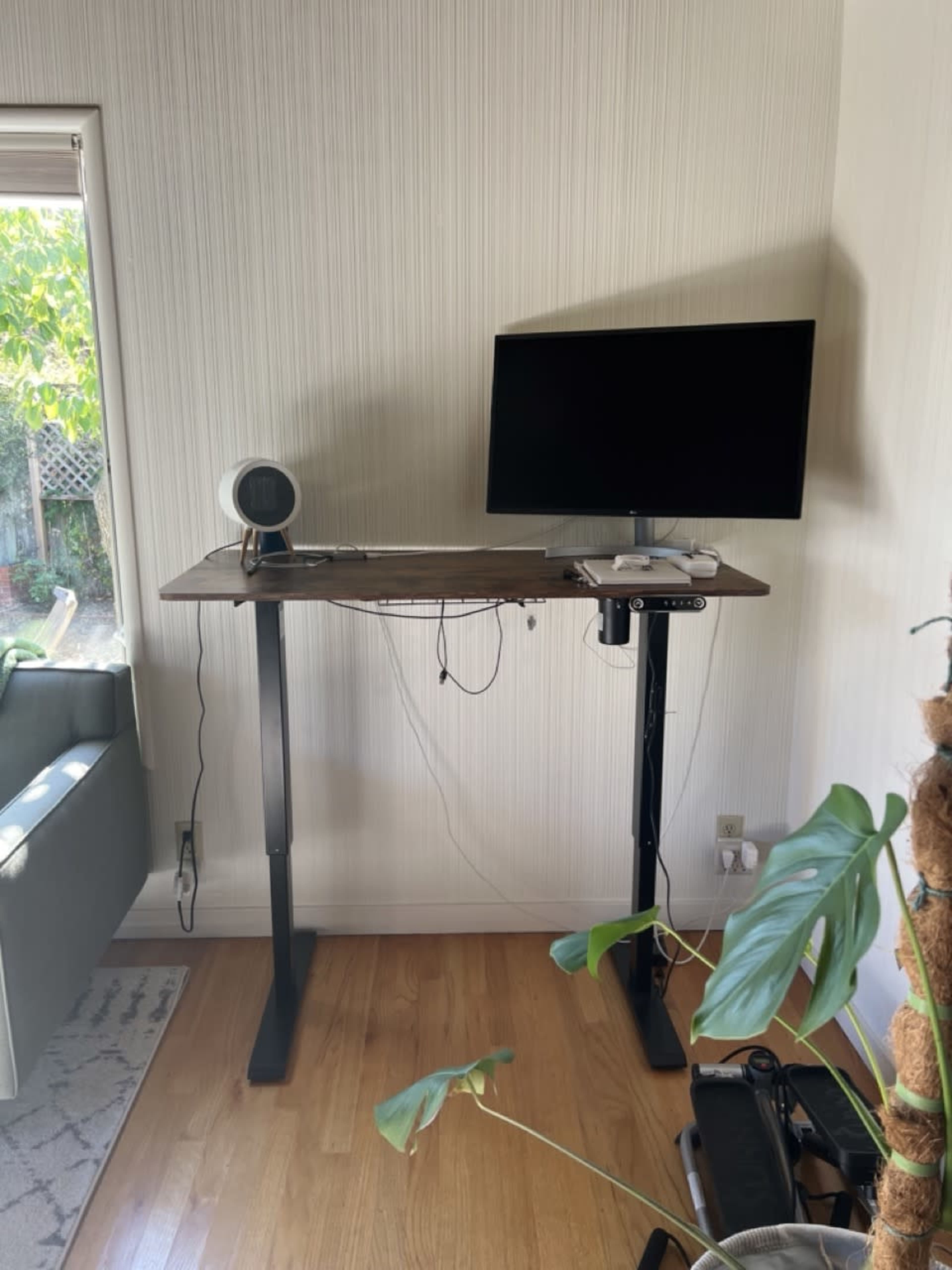 A standing desk with a computer monitor and a lamp is positioned against a wall in a room with wooden flooring.