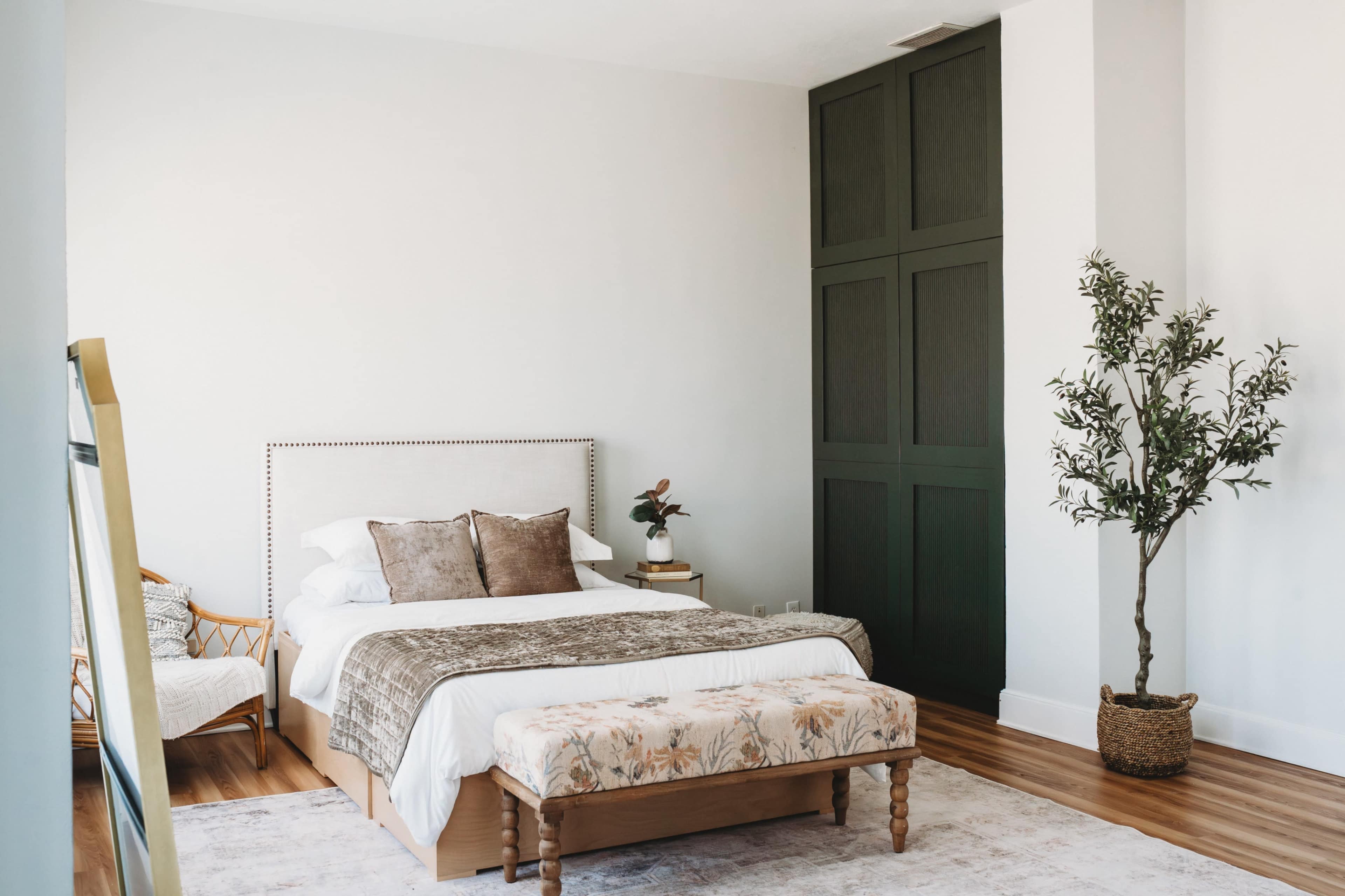 The image shows a modern bedroom featuring a bed with a light-colored headboard, a decorative bench at the foot, and a potted plant in the corner.