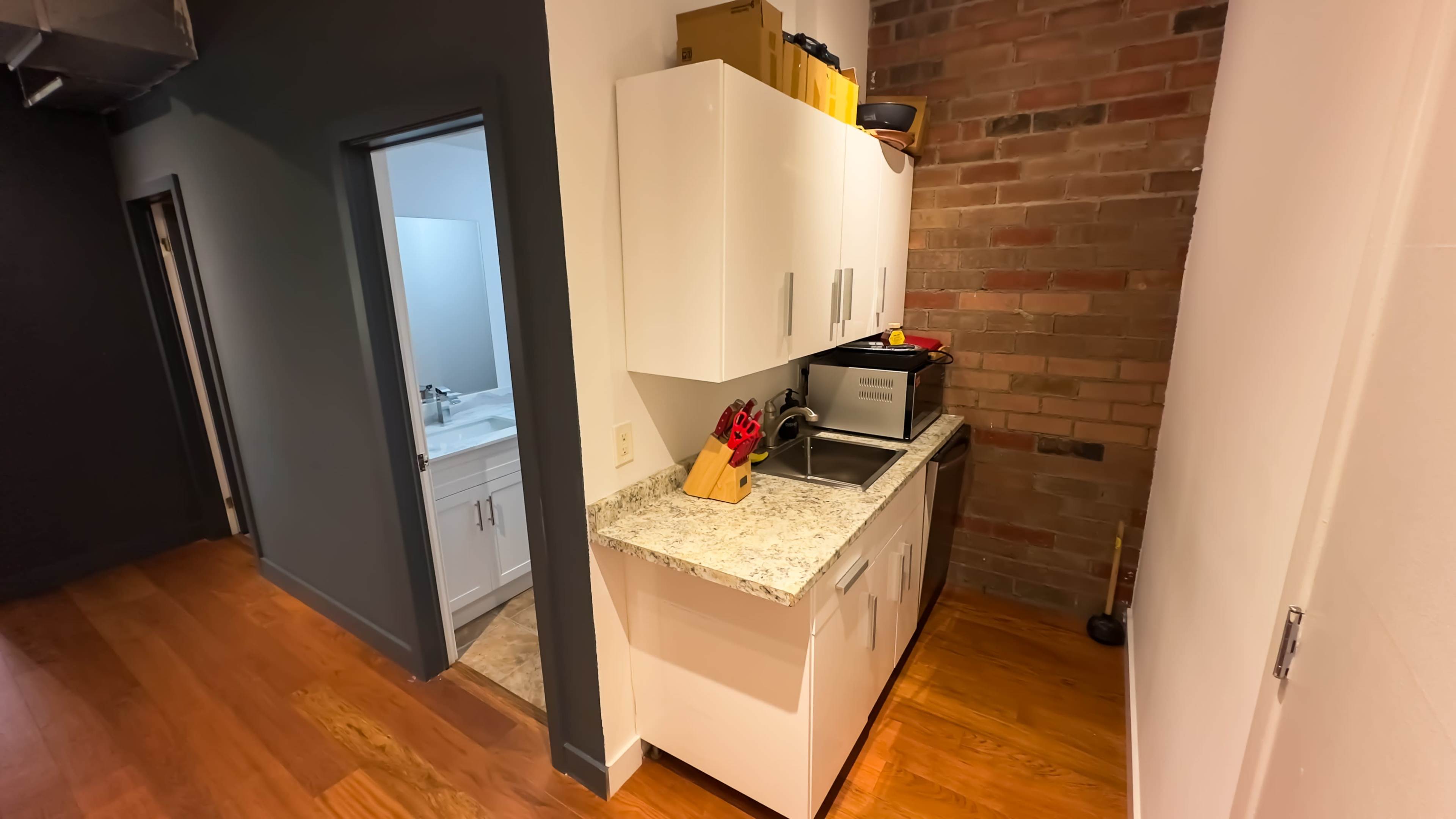 The image shows a compact kitchen area with white cabinets, a sink, and a countertop, adjacent to a doorway leading to a bathroom.
