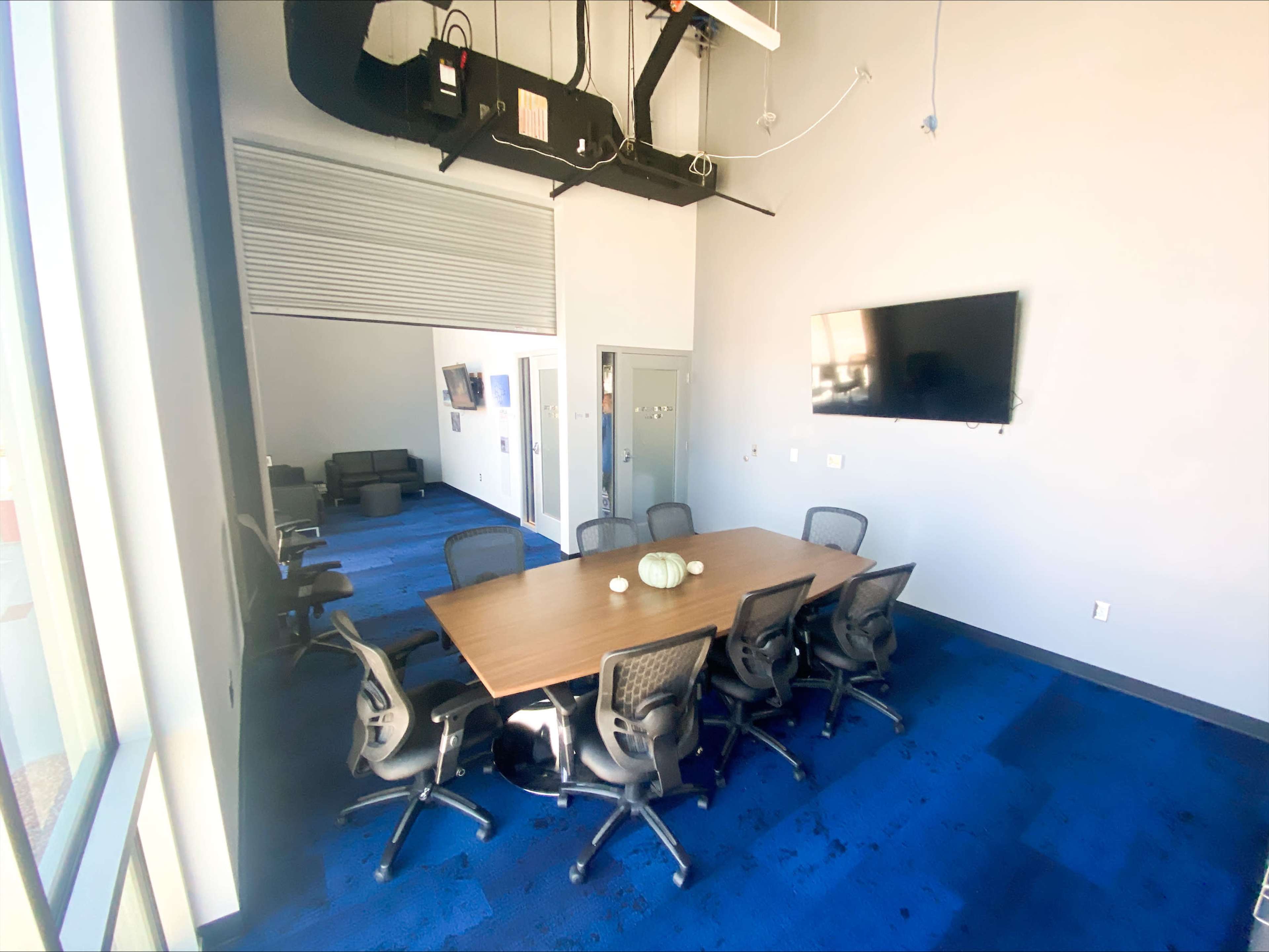 A modern conference room features a large wooden table surrounded by ergonomic chairs, with a wall-mounted screen and a couch visible in the background.