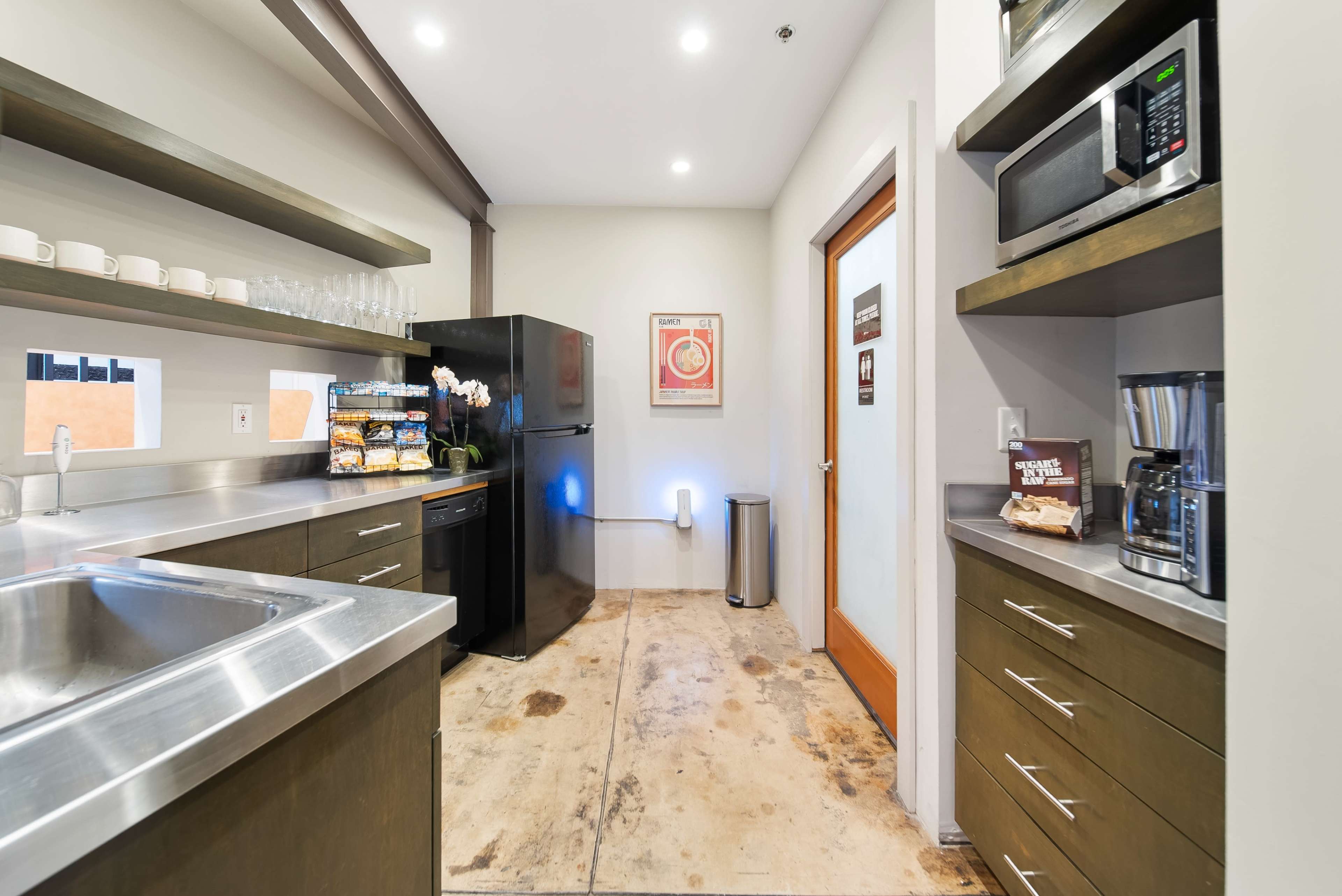 A kitchen area features stainless steel countertops, a black refrigerator, and various appliances, with cabinets and a door leading to another room.