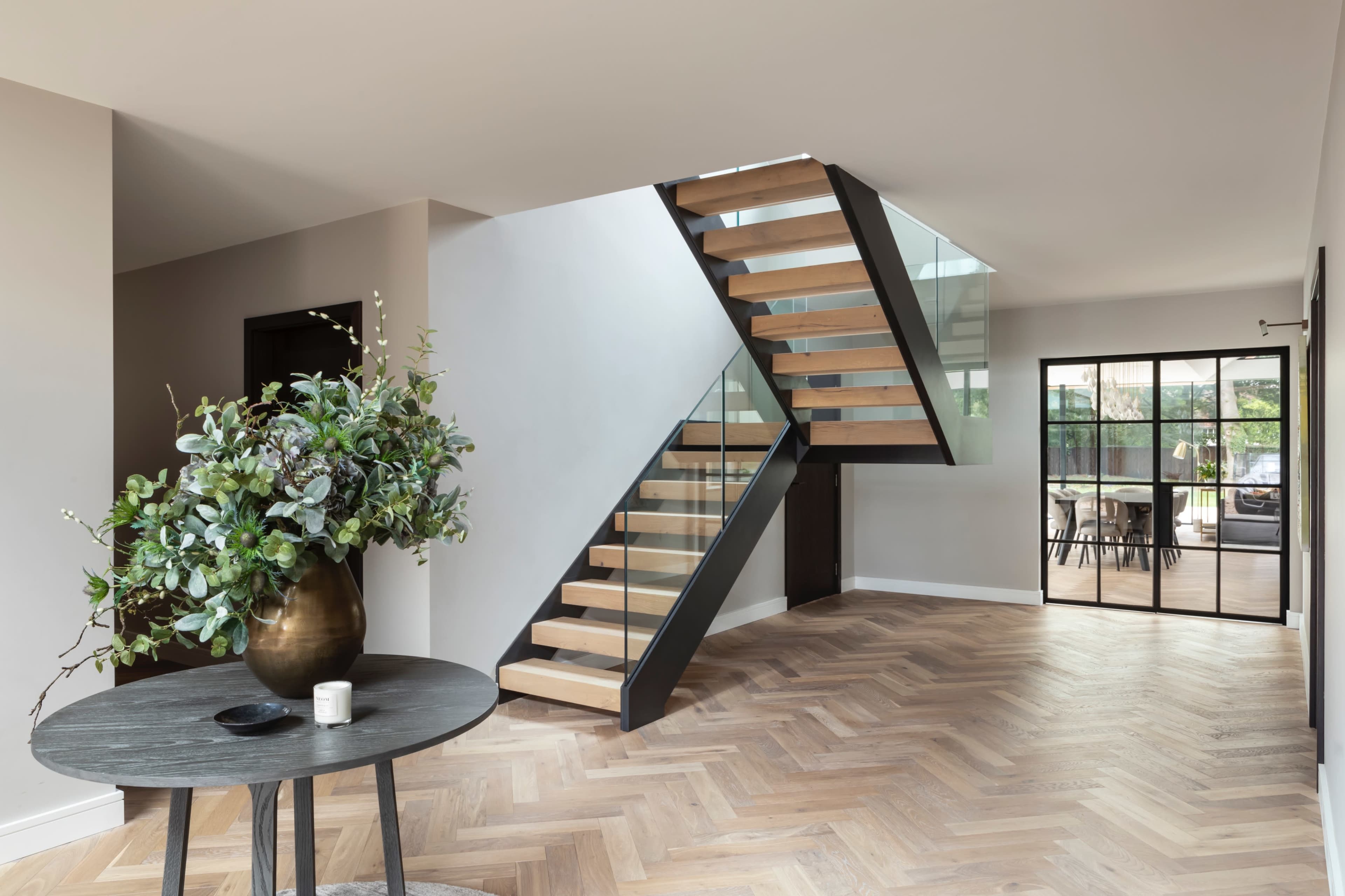 The image shows a contemporary interior with a wooden staircase, a round table with a vase of greenery, and a view of glass doors leading to another room.