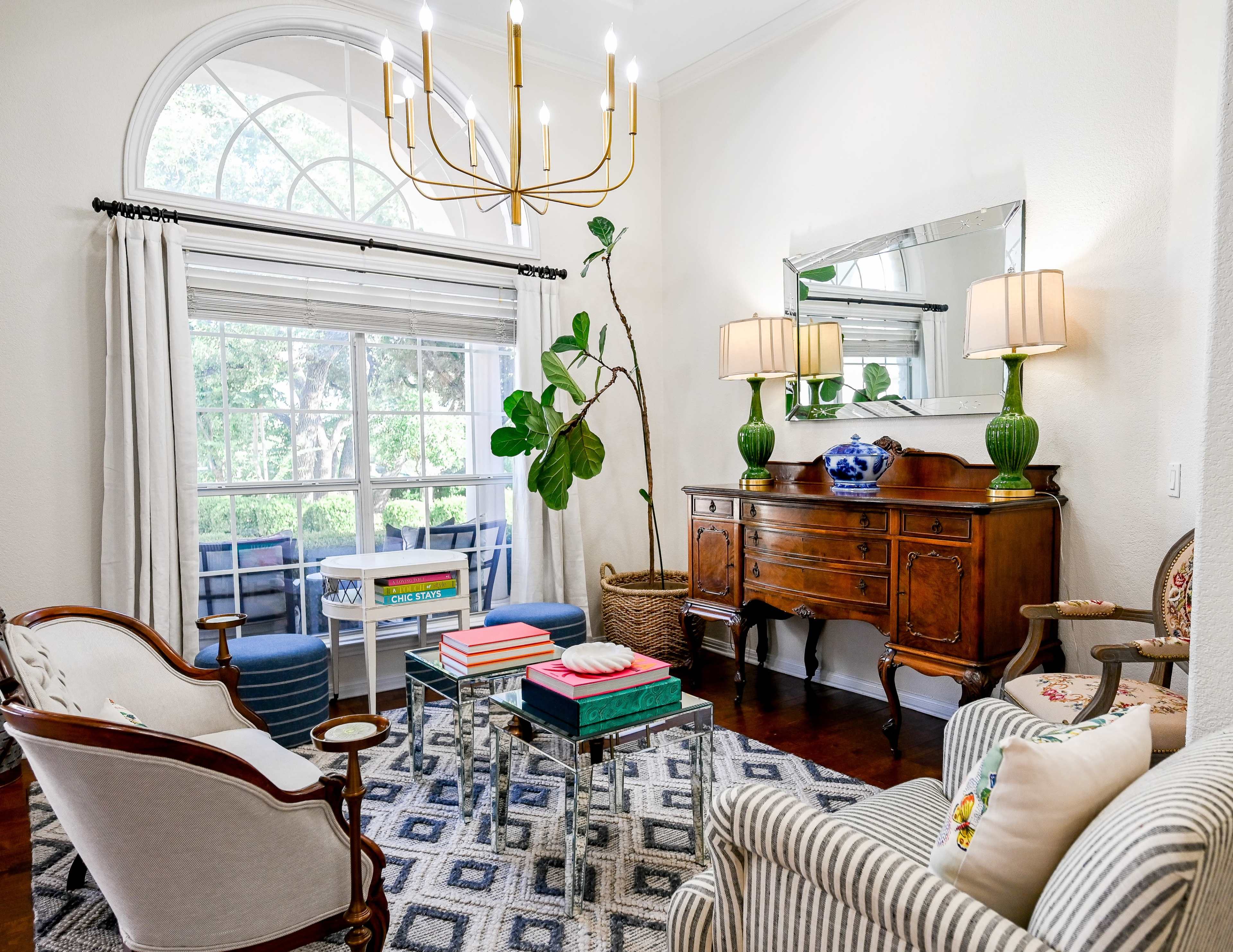 The image shows a well-furnished living room featuring a mix of vintage and modern furniture, a large window with natural light, and decorative plants.