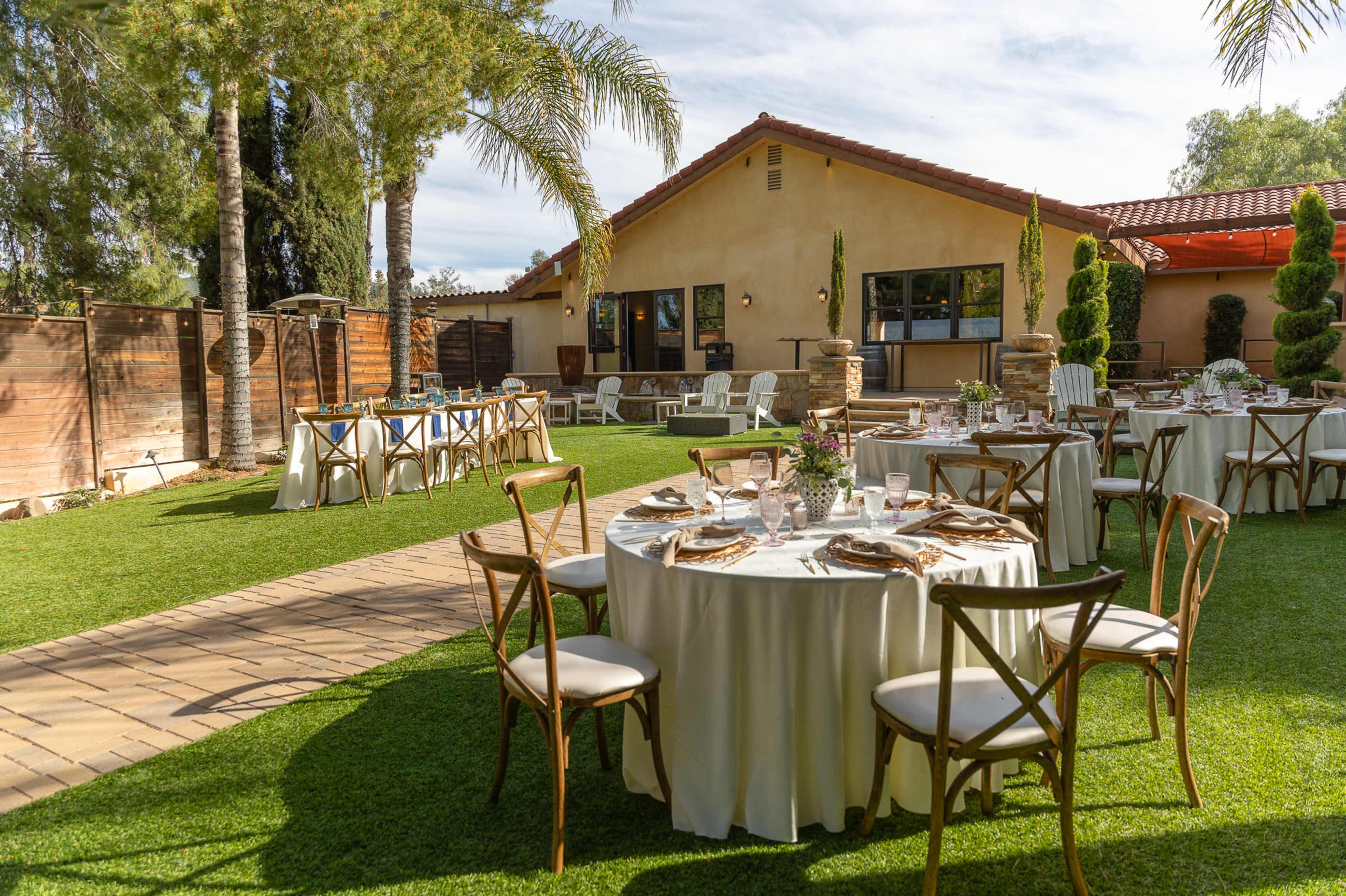 The image shows a well-decorated outdoor space with several round tables set for a dining event, surrounded by greenery and a building in the background.