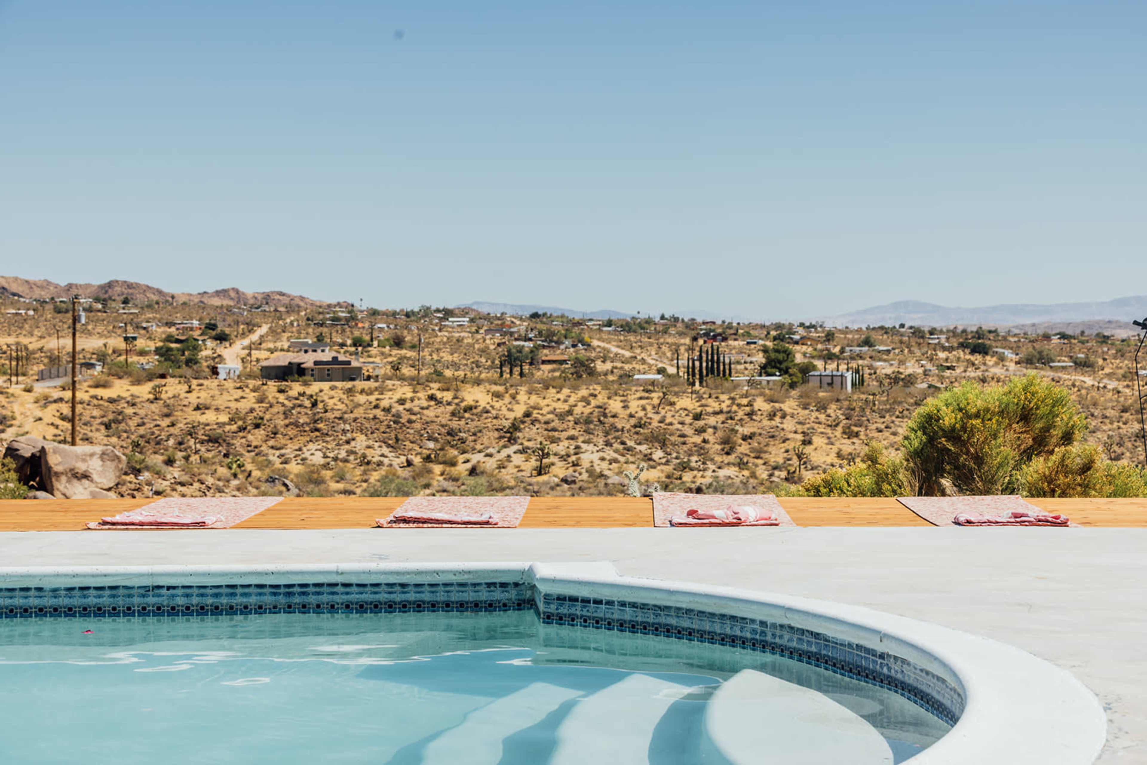 The image shows a swimming pool with a view of arid, desert landscape and sparse buildings in the background.