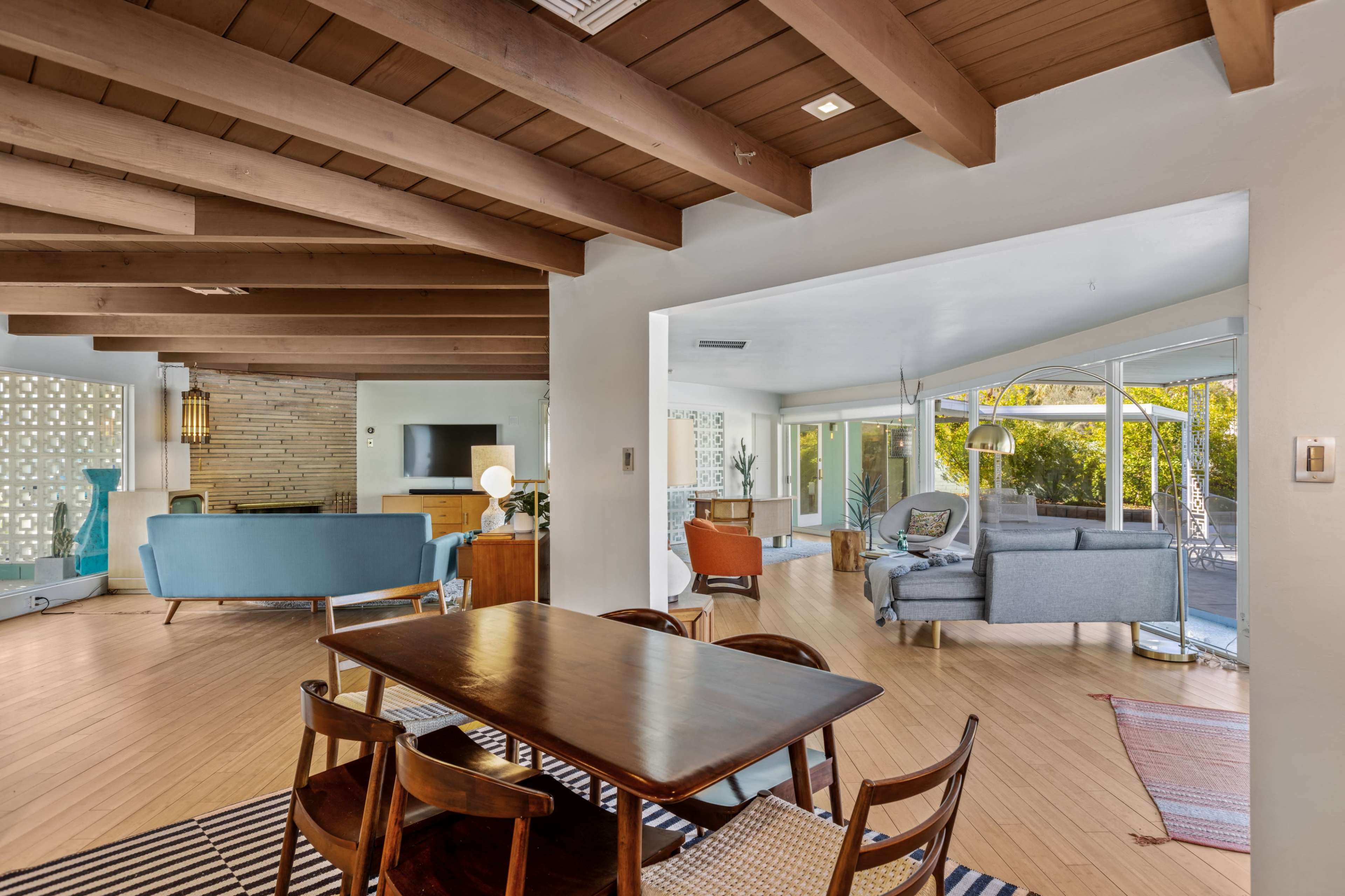 A modern interior space featuring a dining area with a wooden table and chairs, a living room with a blue sofa, and large windows that open to an outdoor area.