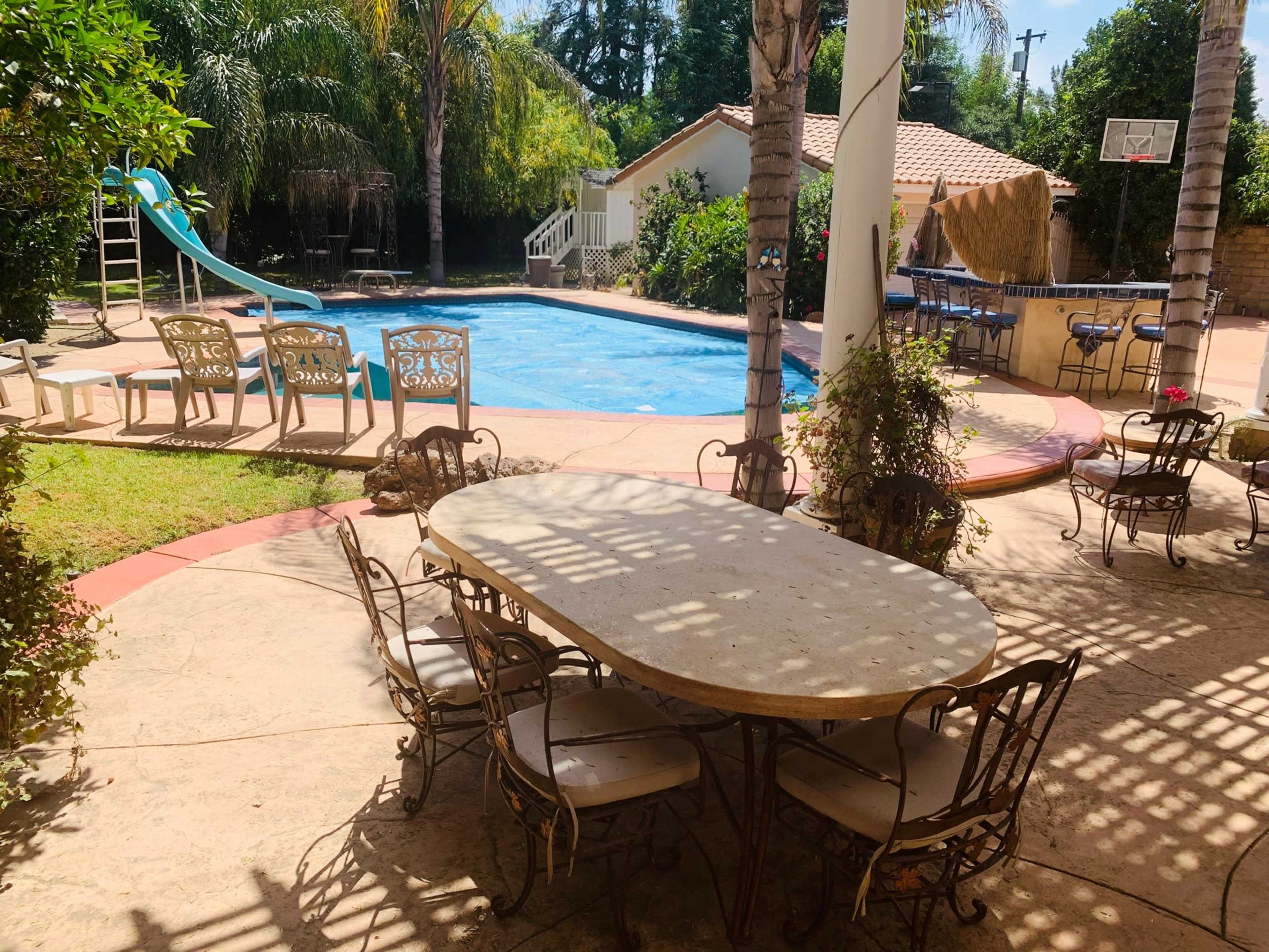 A backyard features a swimming pool surrounded by palm trees, lounge chairs, and a dining table set for several people.
