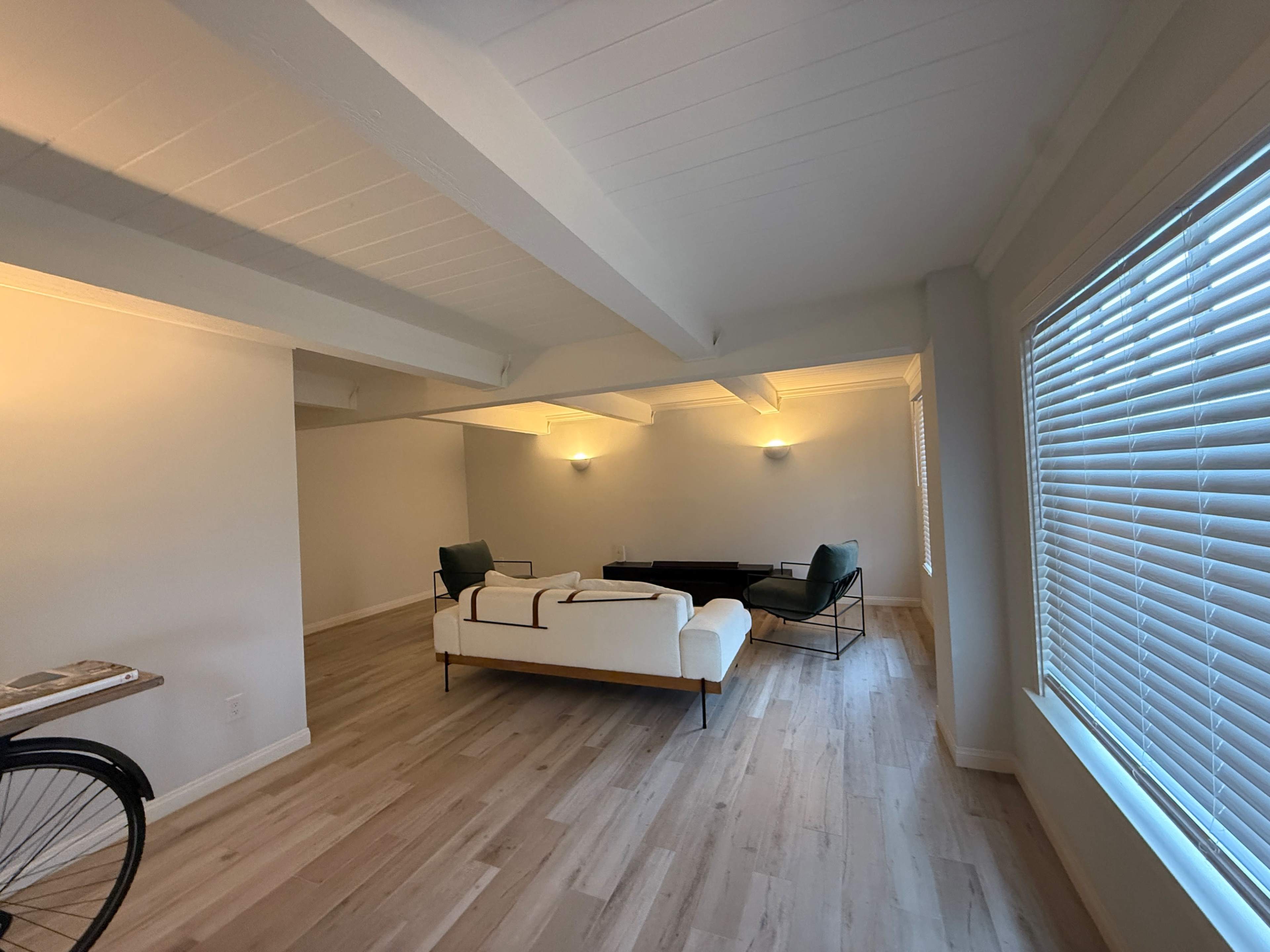 The image shows a spacious, minimally furnished living area with a white couch and dark chairs arranged under modern lighting.