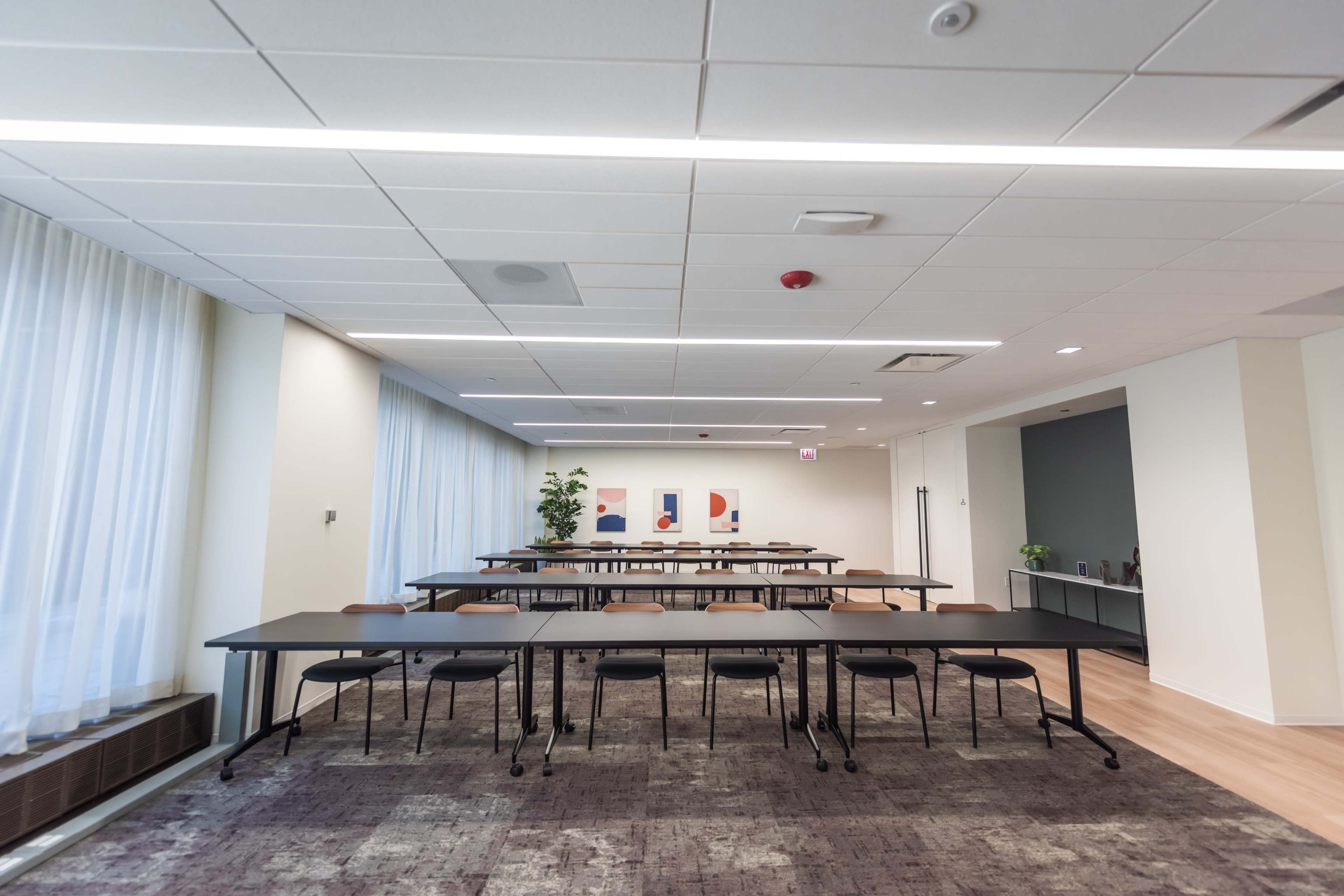 A modern conference room features several long tables arranged in a U-shape, with minimalist decor and large windows allowing natural light.