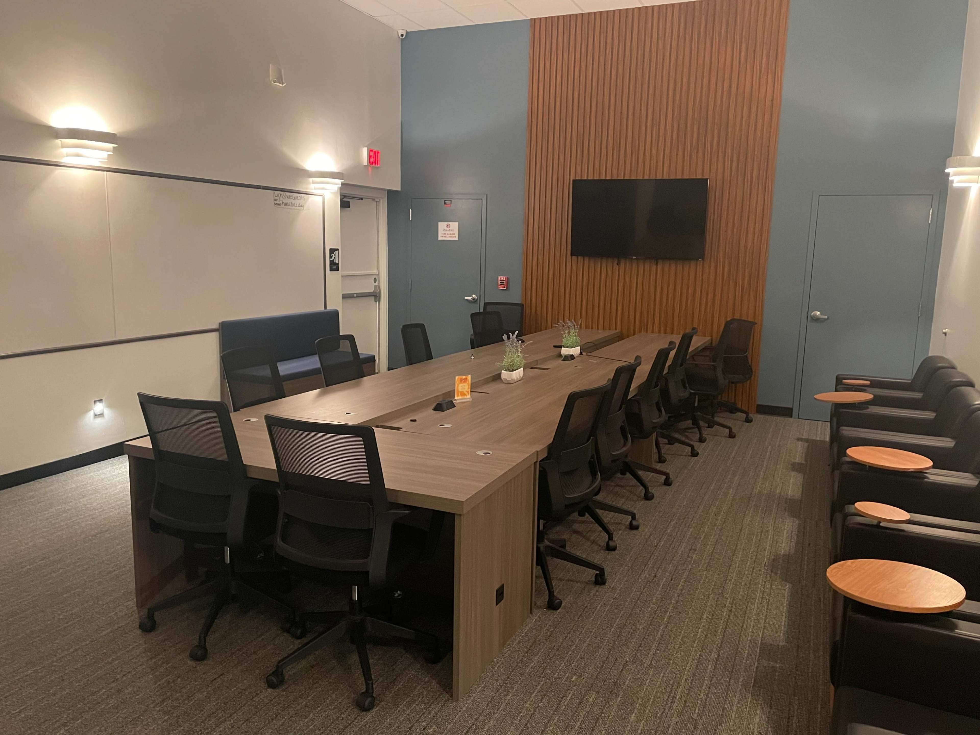 A conference room features a long wooden table surrounded by black chairs, with a television on the wall and plants on the table.