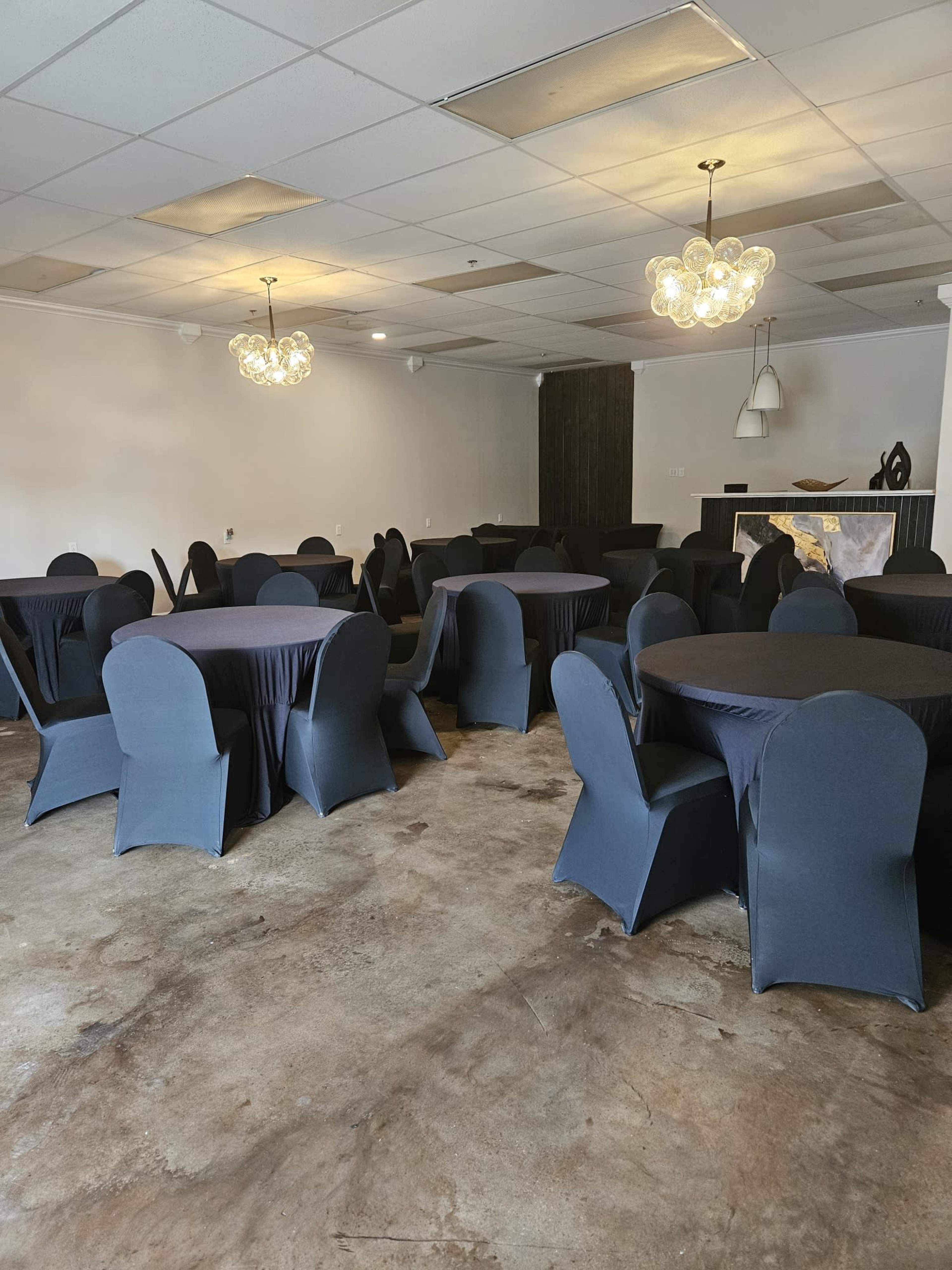 The image shows a banquet room set up with round tables covered in navy tablecloths and dark chairs, featuring pendant lighting and a decorative wall.