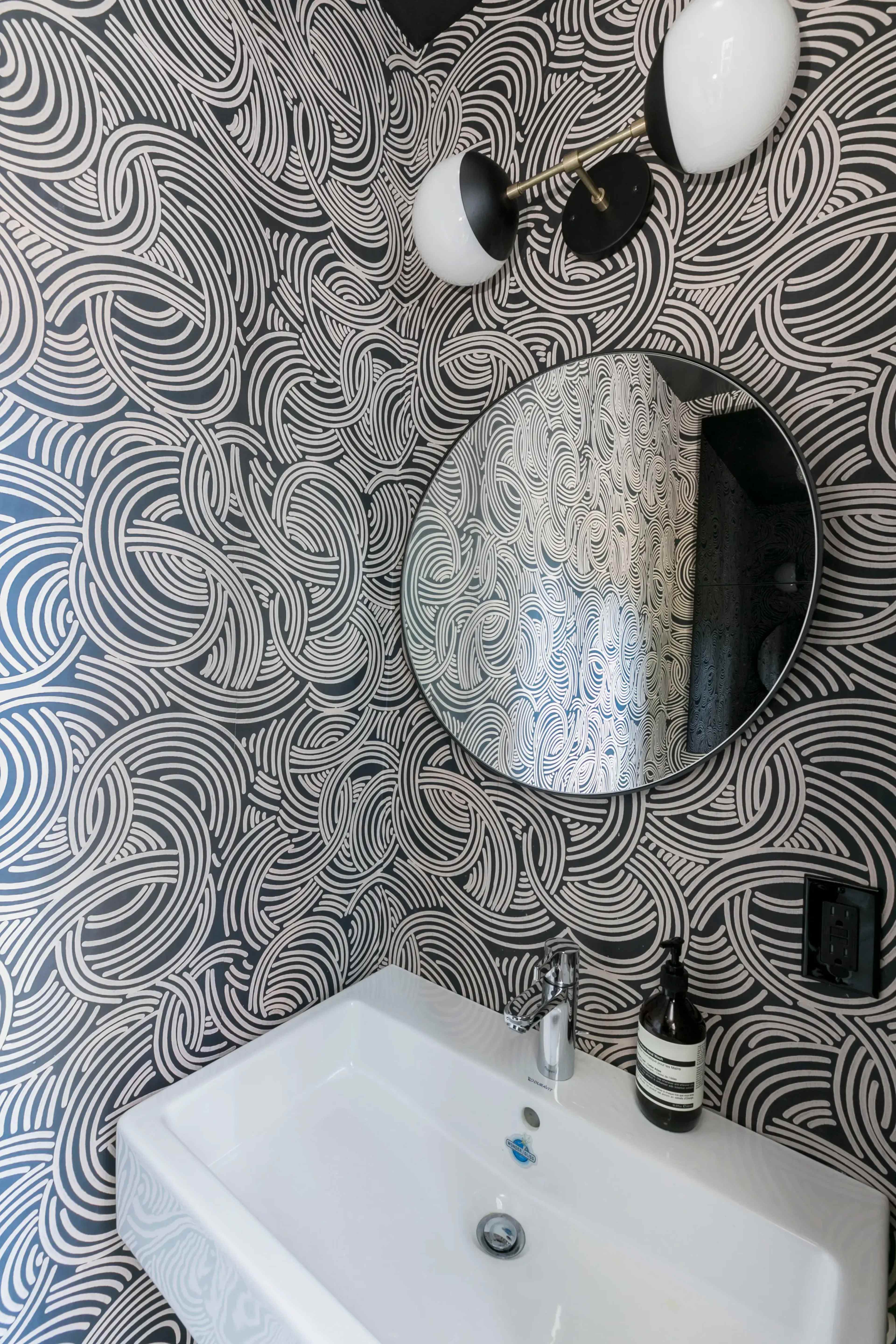 A modern bathroom features a circular mirror above a sleek white sink against a stylized black and white swirl-patterned wall.