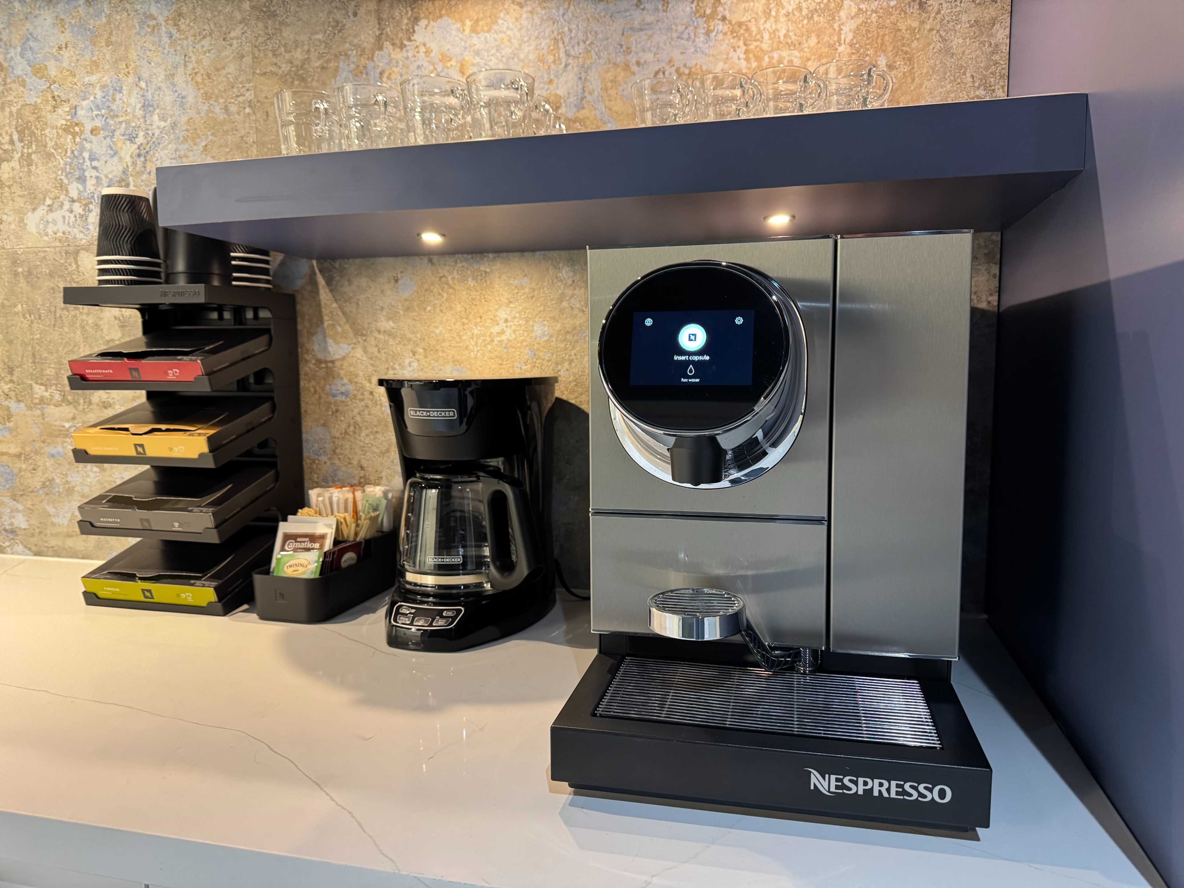 The image shows a modern coffee station featuring a Nespresso machine, a coffee maker, and a shelf with glassware and organizational bins.