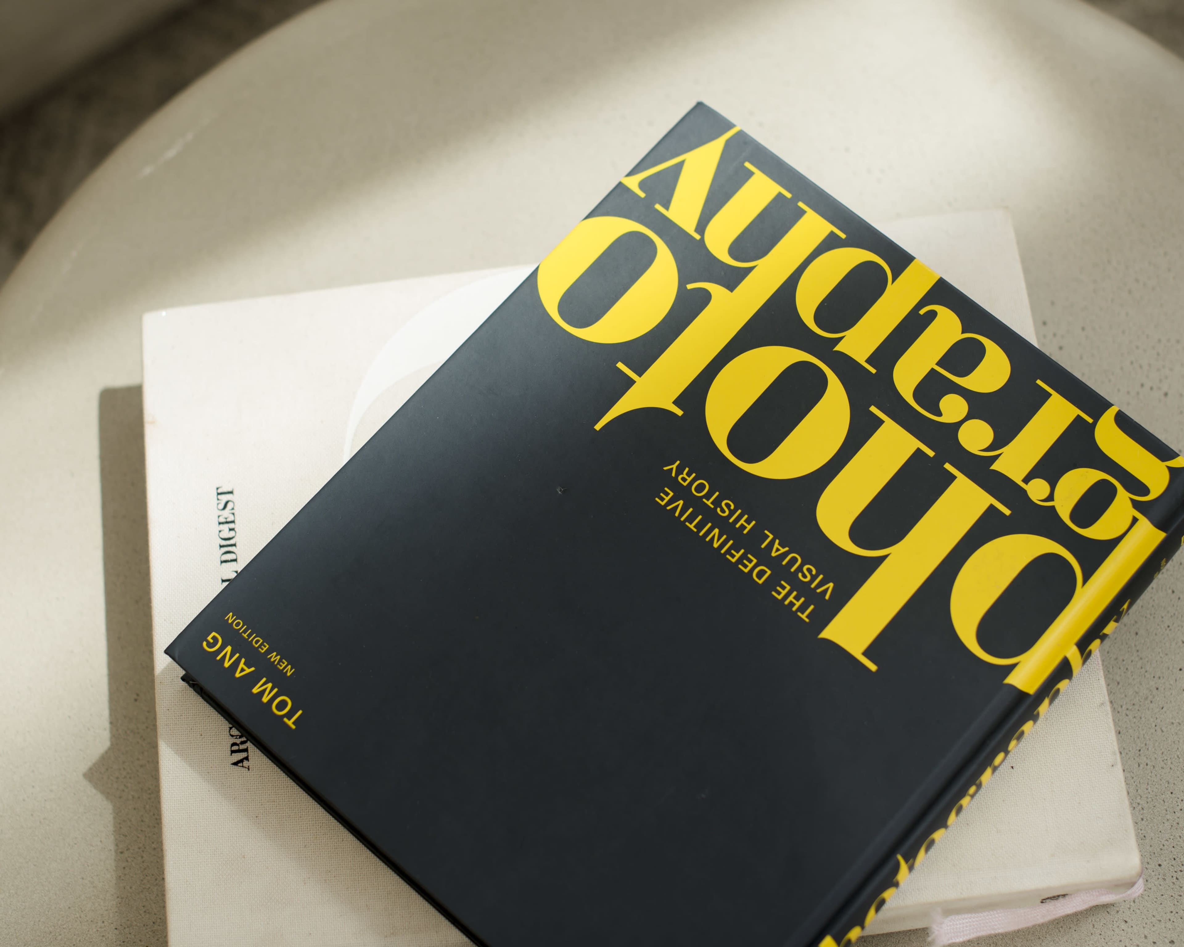A black book titled "Photography: The Definitive Visual History" by Tom Ang is positioned upside down on a white surface beside another lighter book.