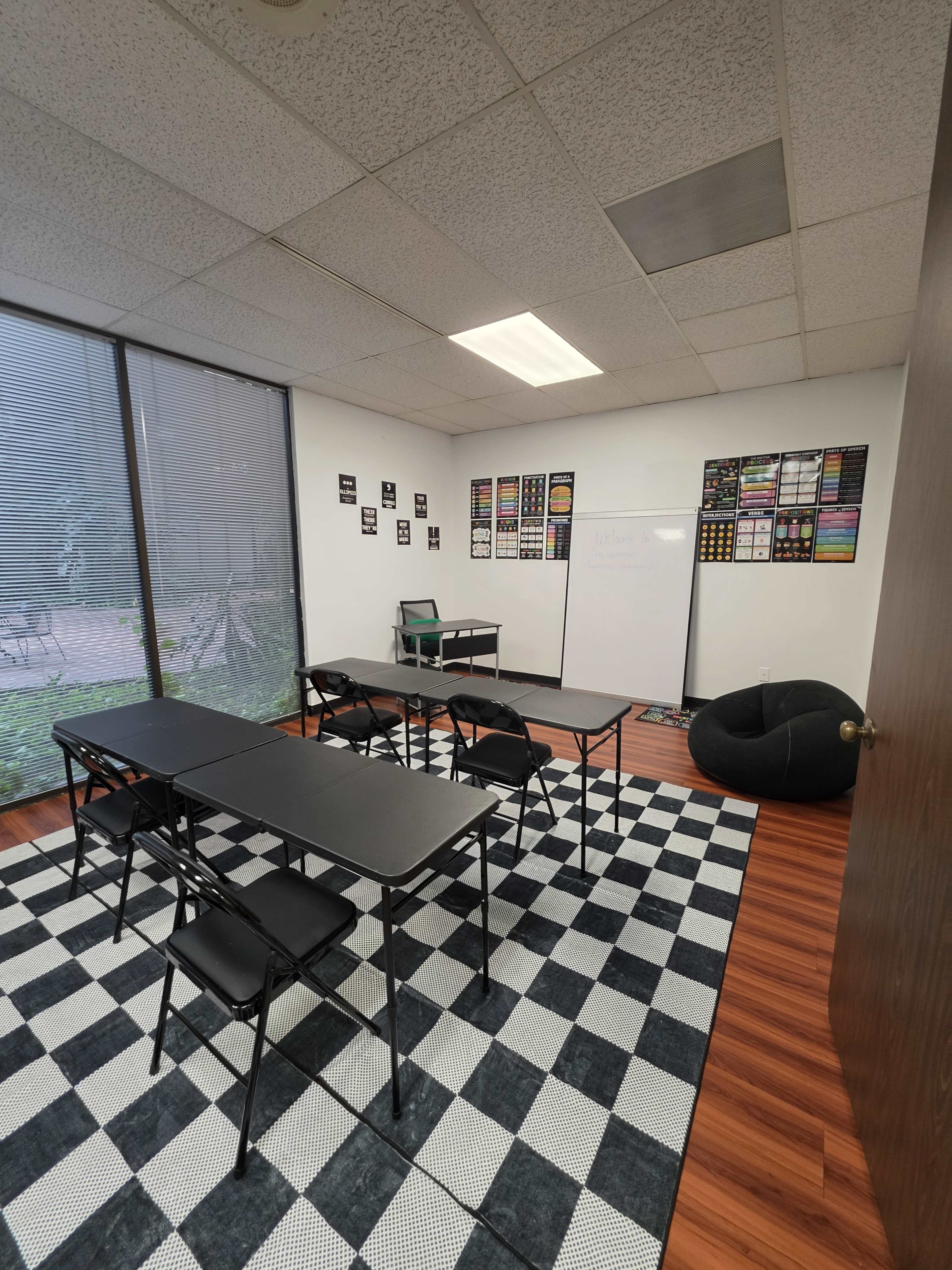 A classroom is set up with black tables and chairs arranged in a grid pattern on a checkered rug, featuring a whiteboard and colorful posters on the walls.