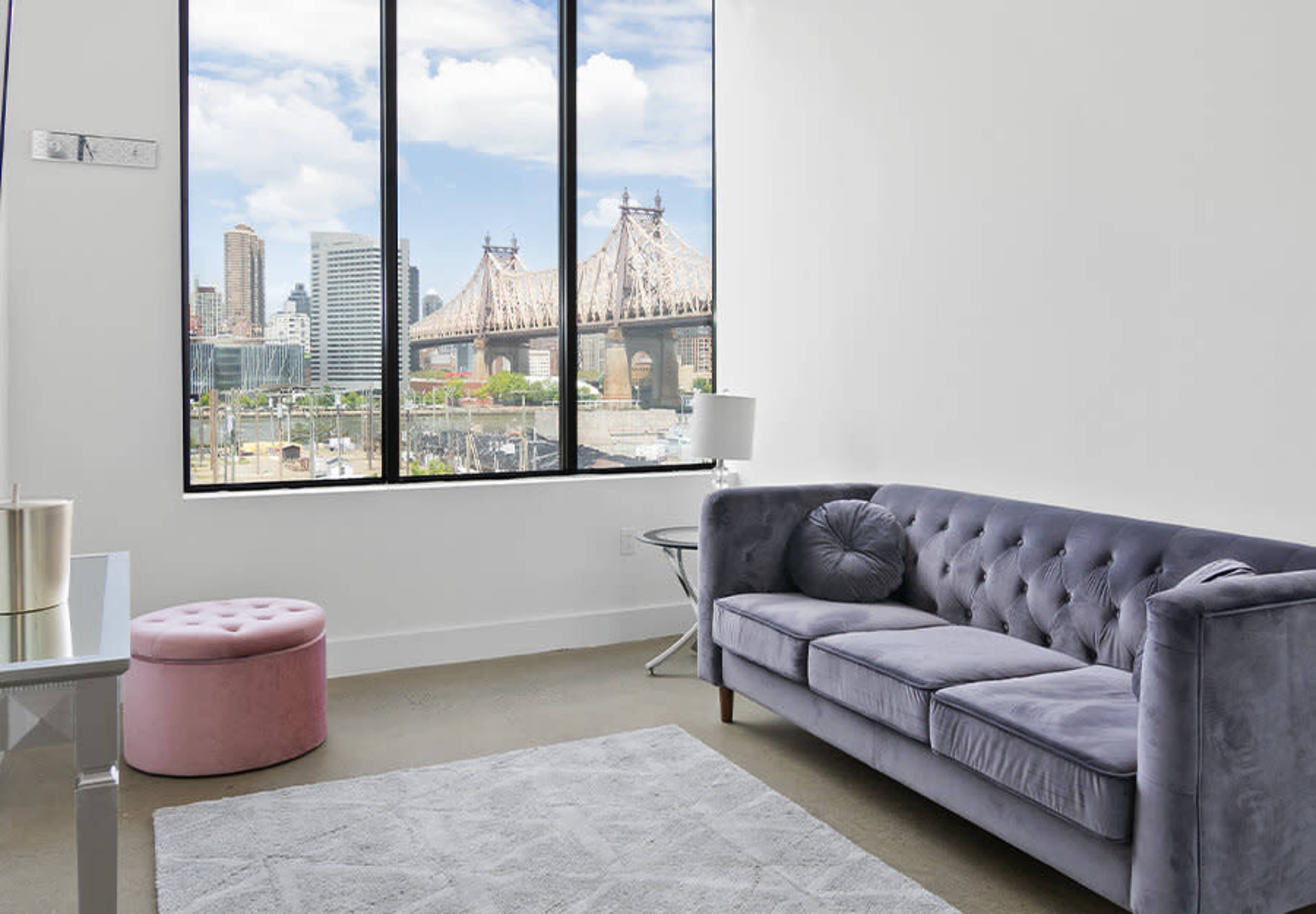 A modern living room features a gray couch and a pink ottoman, with large windows displaying a view of city buildings and a bridge.