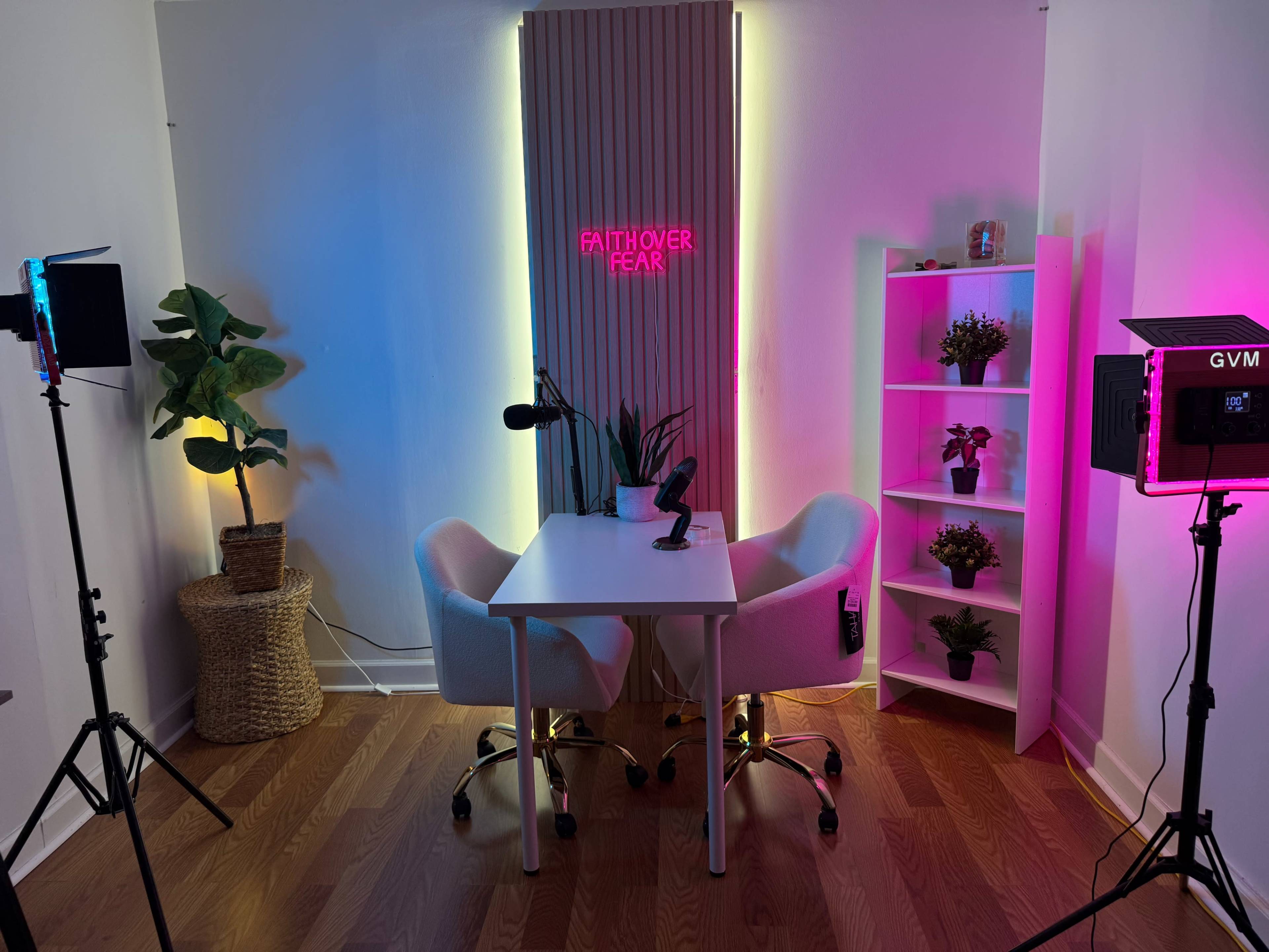 The image shows a brightly lit podcast studio featuring a white table, two chairs, a neon sign, and various plants arranged around the space.
