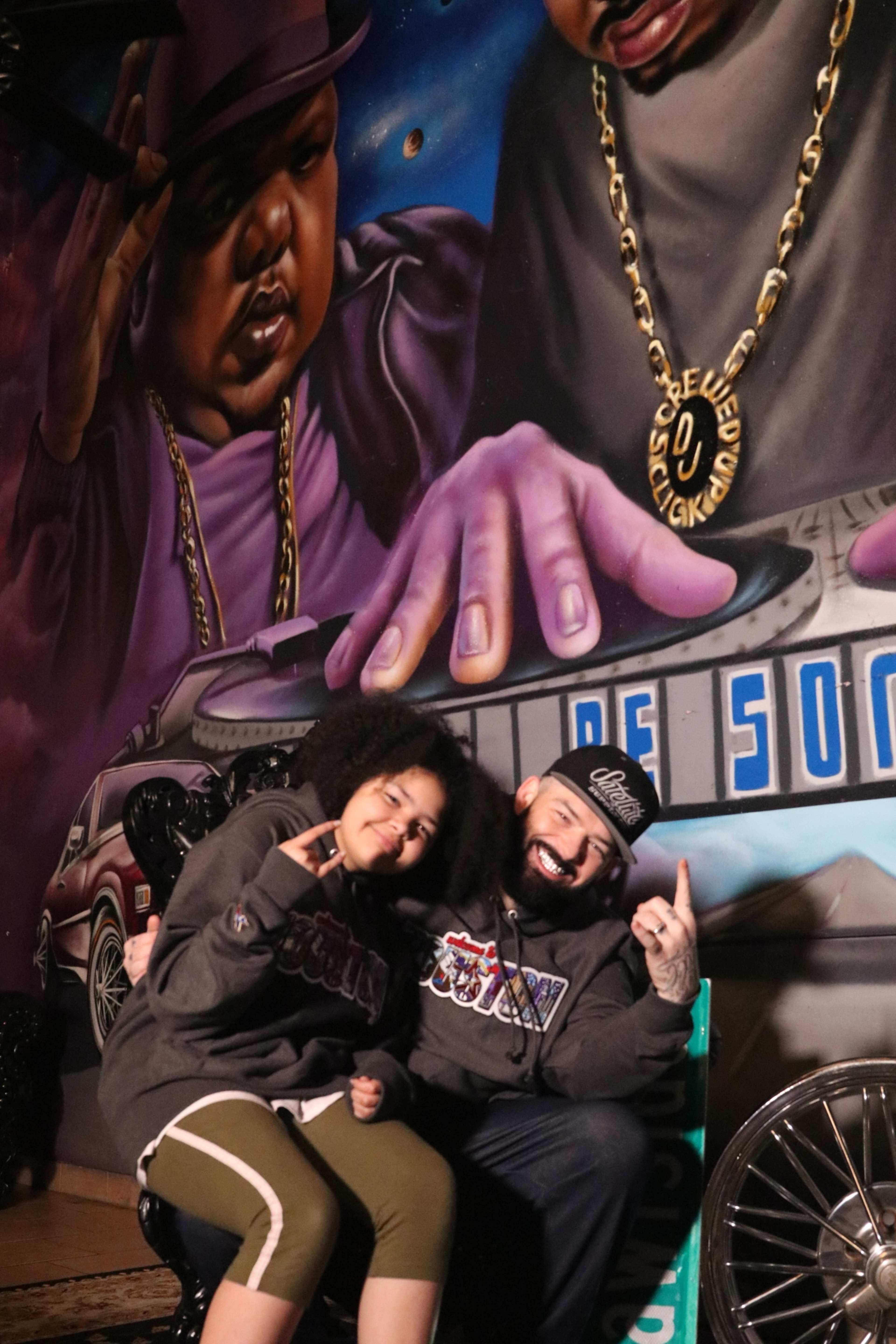 A man and a girl sit in front of a colorful mural featuring notable hip-hop figures.