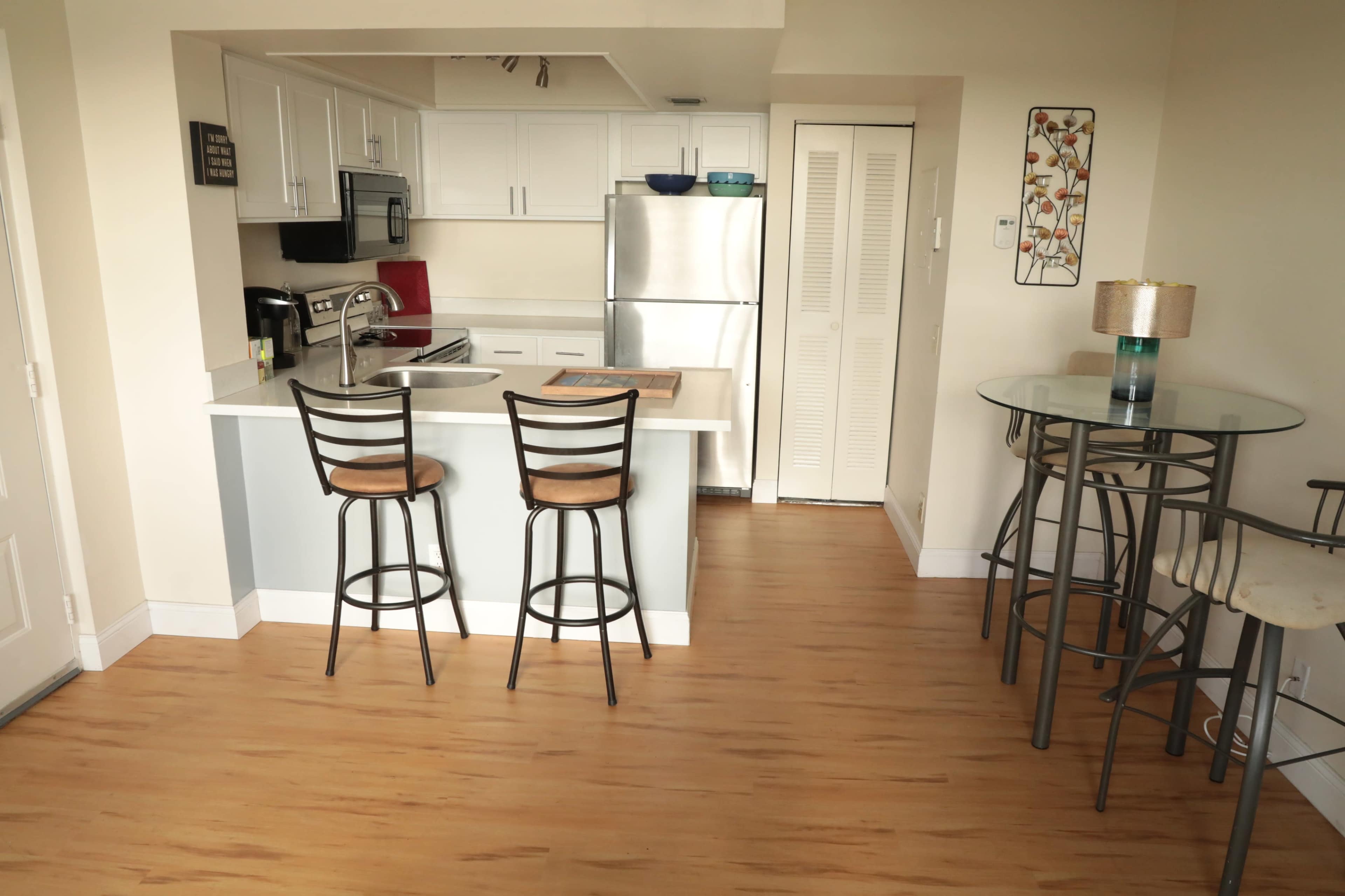 A modern kitchen features white cabinets, stainless steel appliances, and a small dining area with two bar stools and a glass-topped table.
