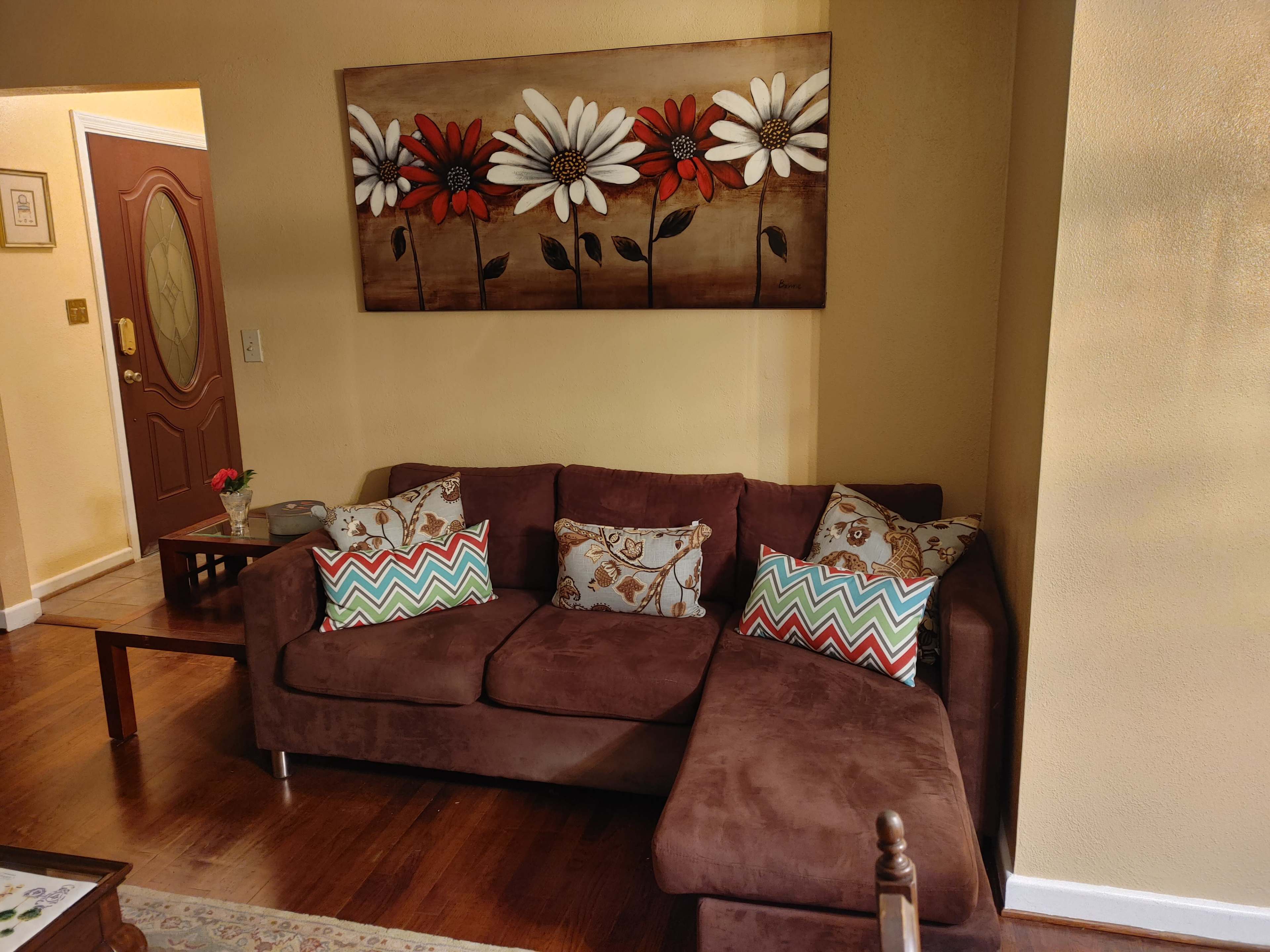 The image shows a cozy living room with a brown sectional sofa adorned with colorful patterned pillows, next to a wooden coffee table and a wall decorated with a painting of flowers.