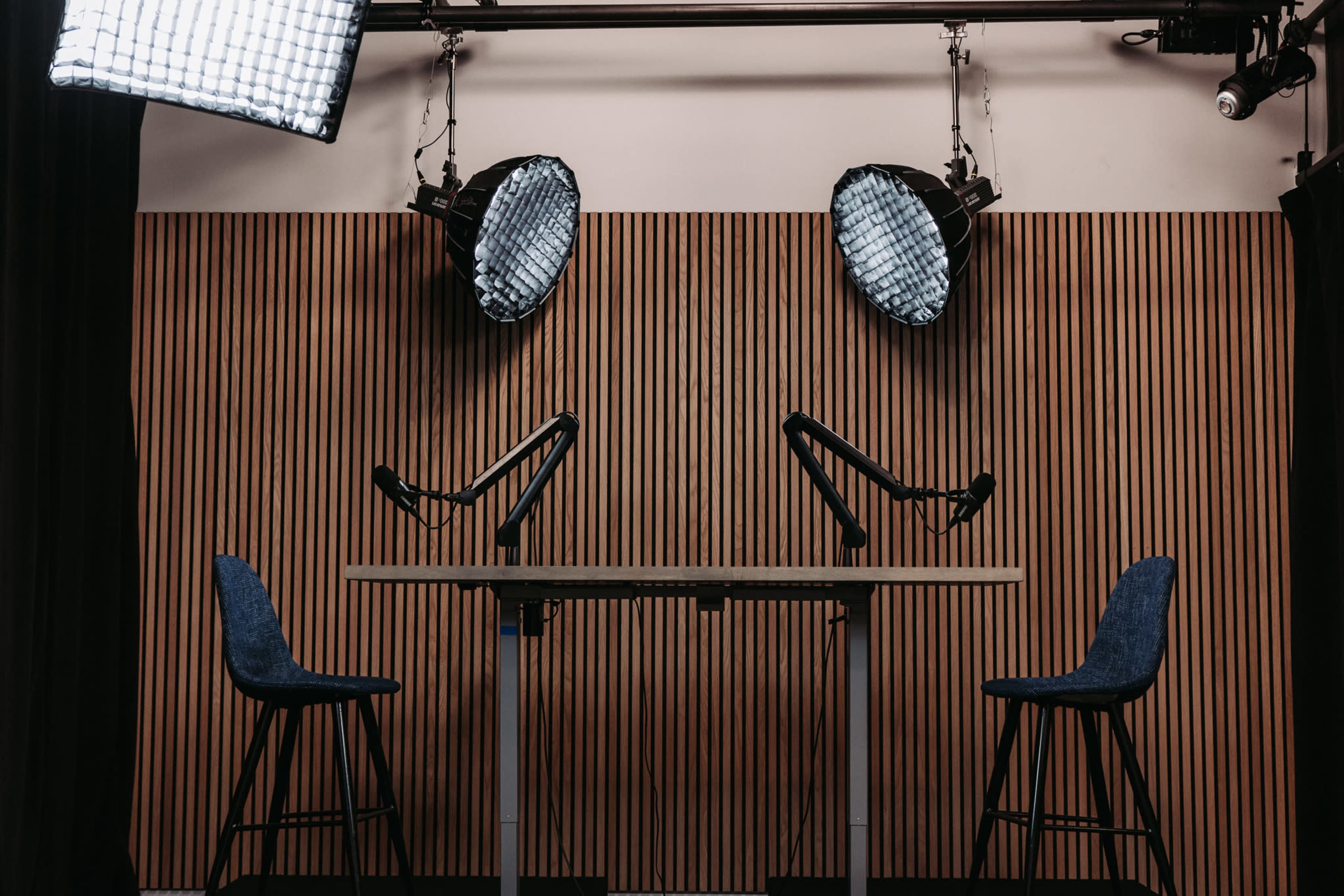 A podcast studio features a wooden wall, two microphone stands, and two chairs under softbox lights.