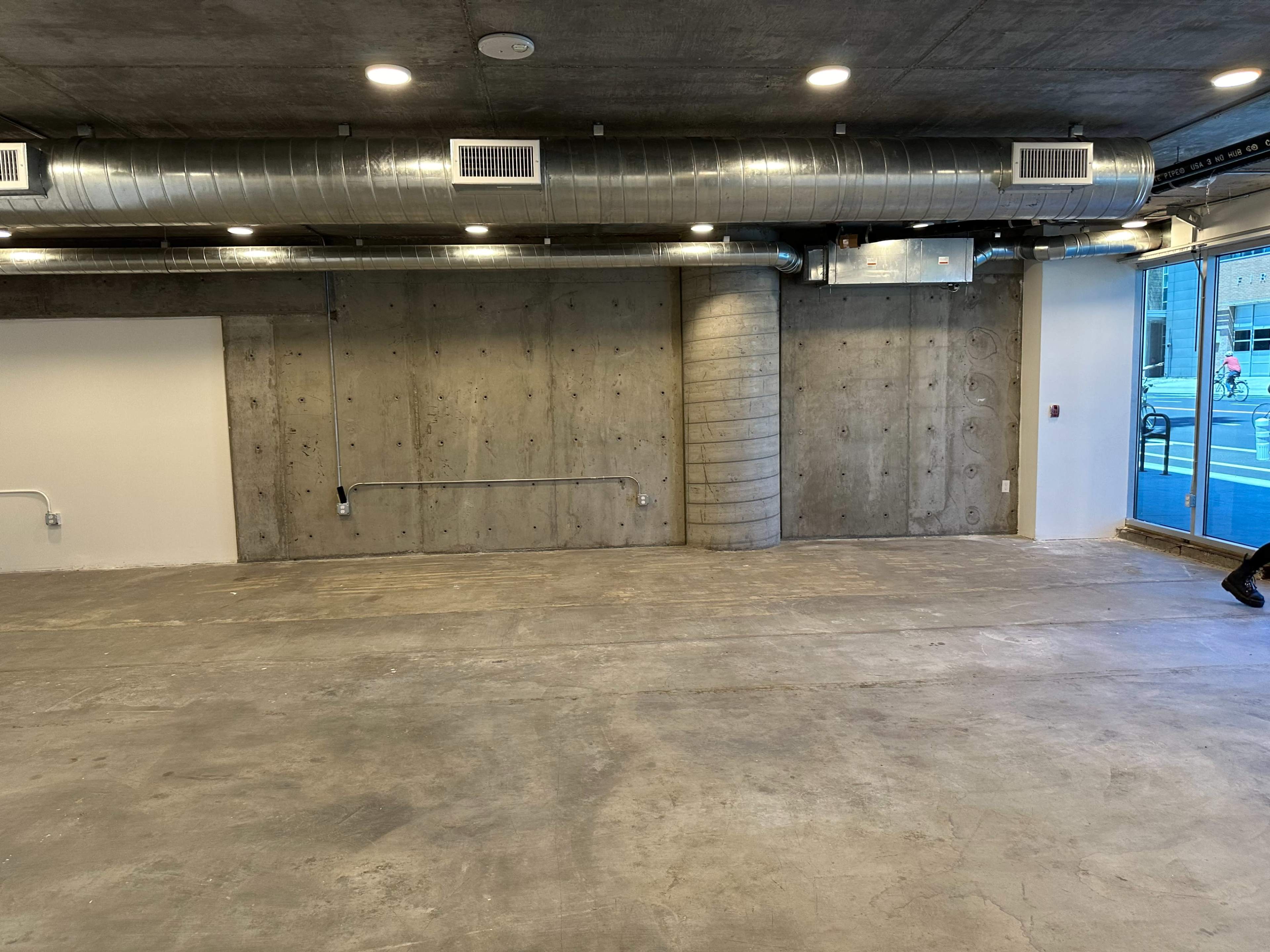 The image shows an empty industrial-style room with concrete walls, exposed ductwork, and a polished concrete floor.