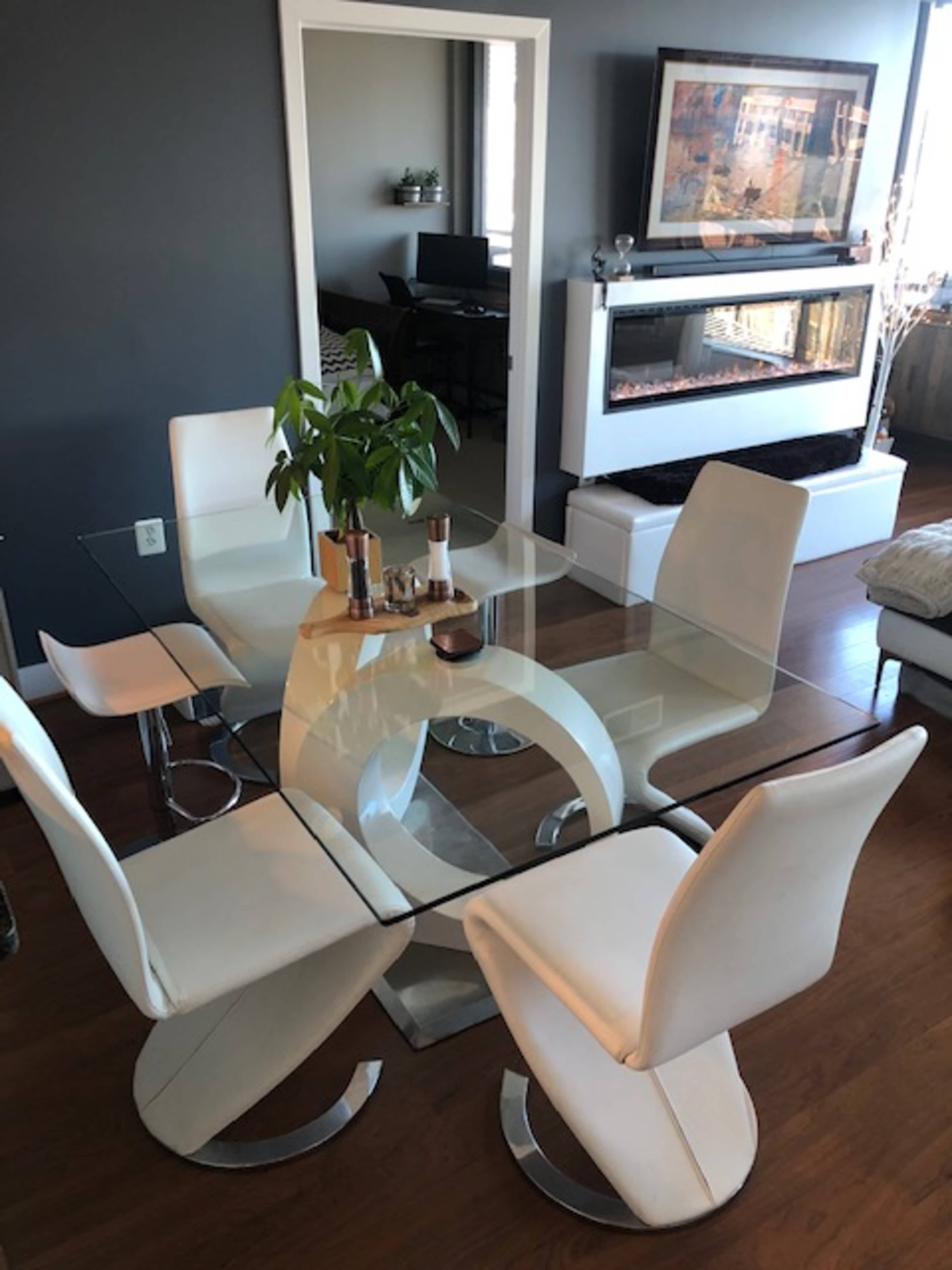 A modern dining area features a glass table with a curved white base surrounded by four white chairs, with an indoor plant in the center.