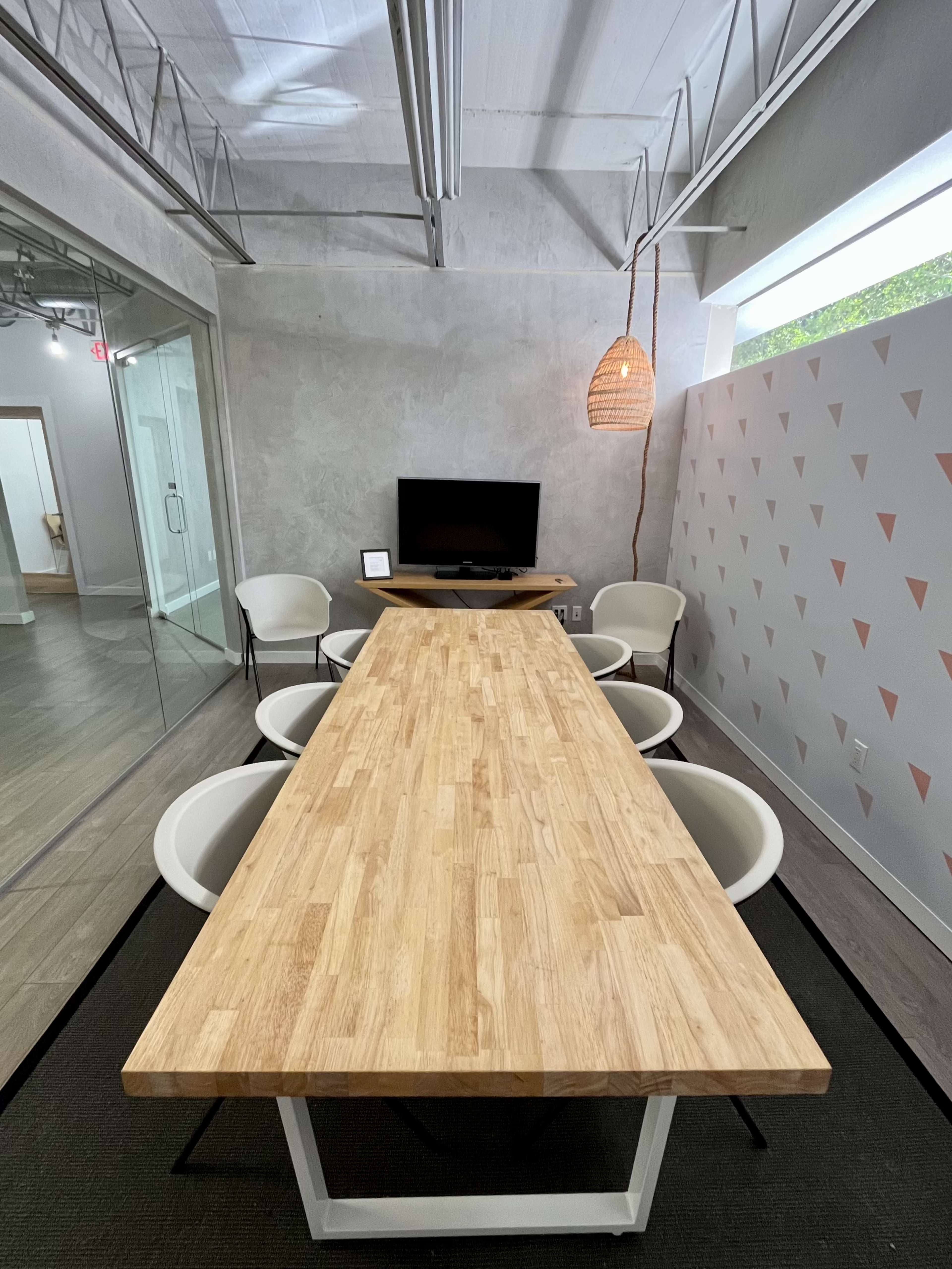 A long wooden table is flanked by four white chairs in a modern conference room with a television and a decorative light fixture.