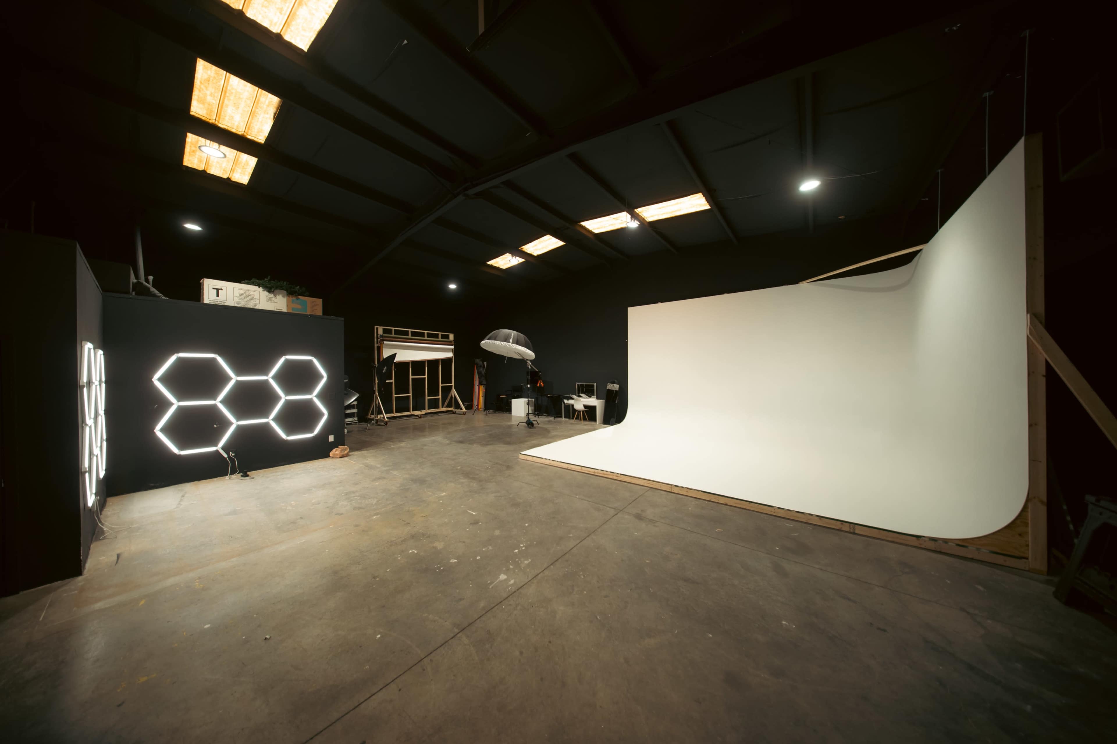 The image shows a spacious studio with a white backdrop on one side and a black wall featuring hexagonal lighting on the other.