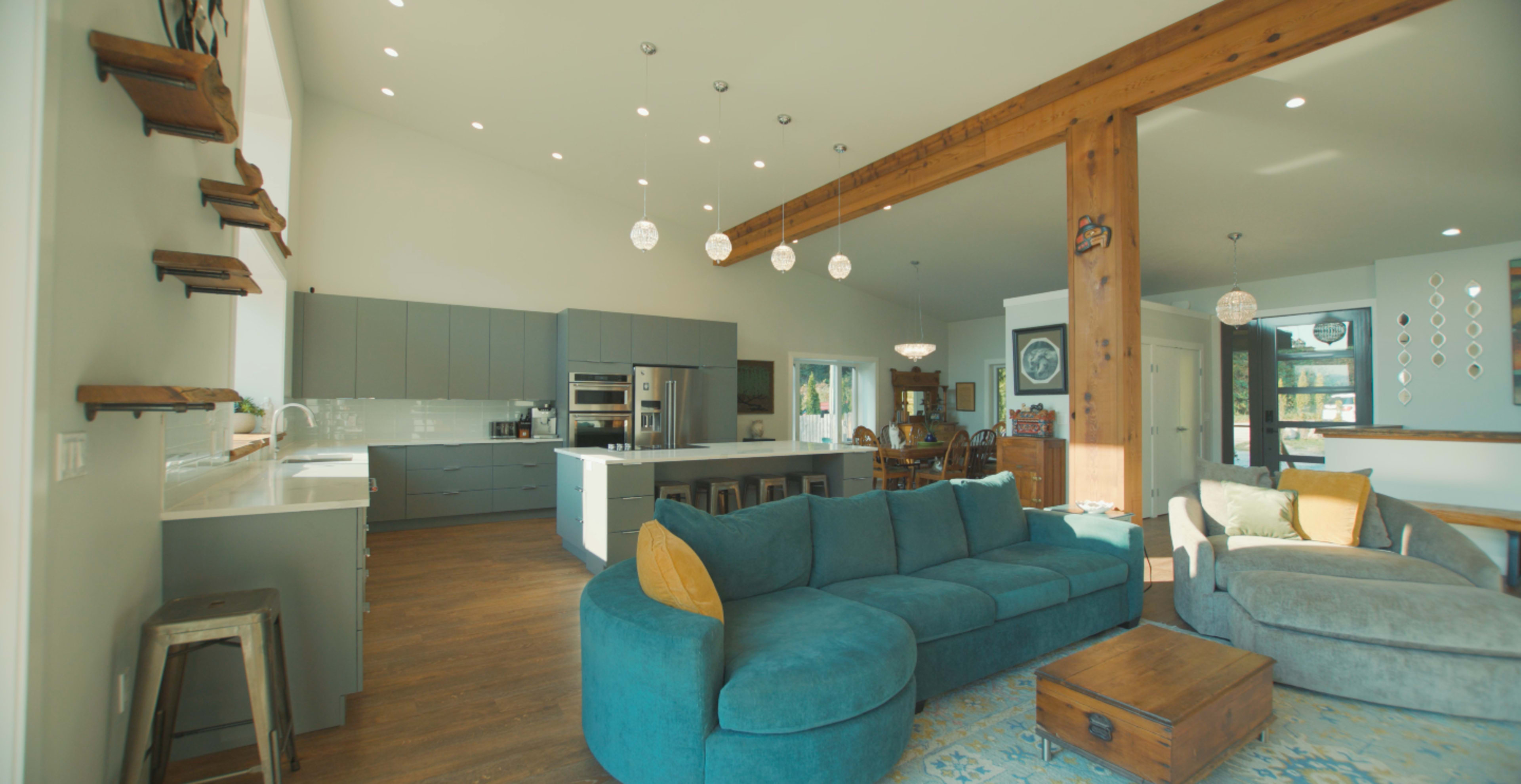 A spacious living area features a large blue sectional sofa, a wooden beam ceiling, and a modern kitchen with gray cabinets.