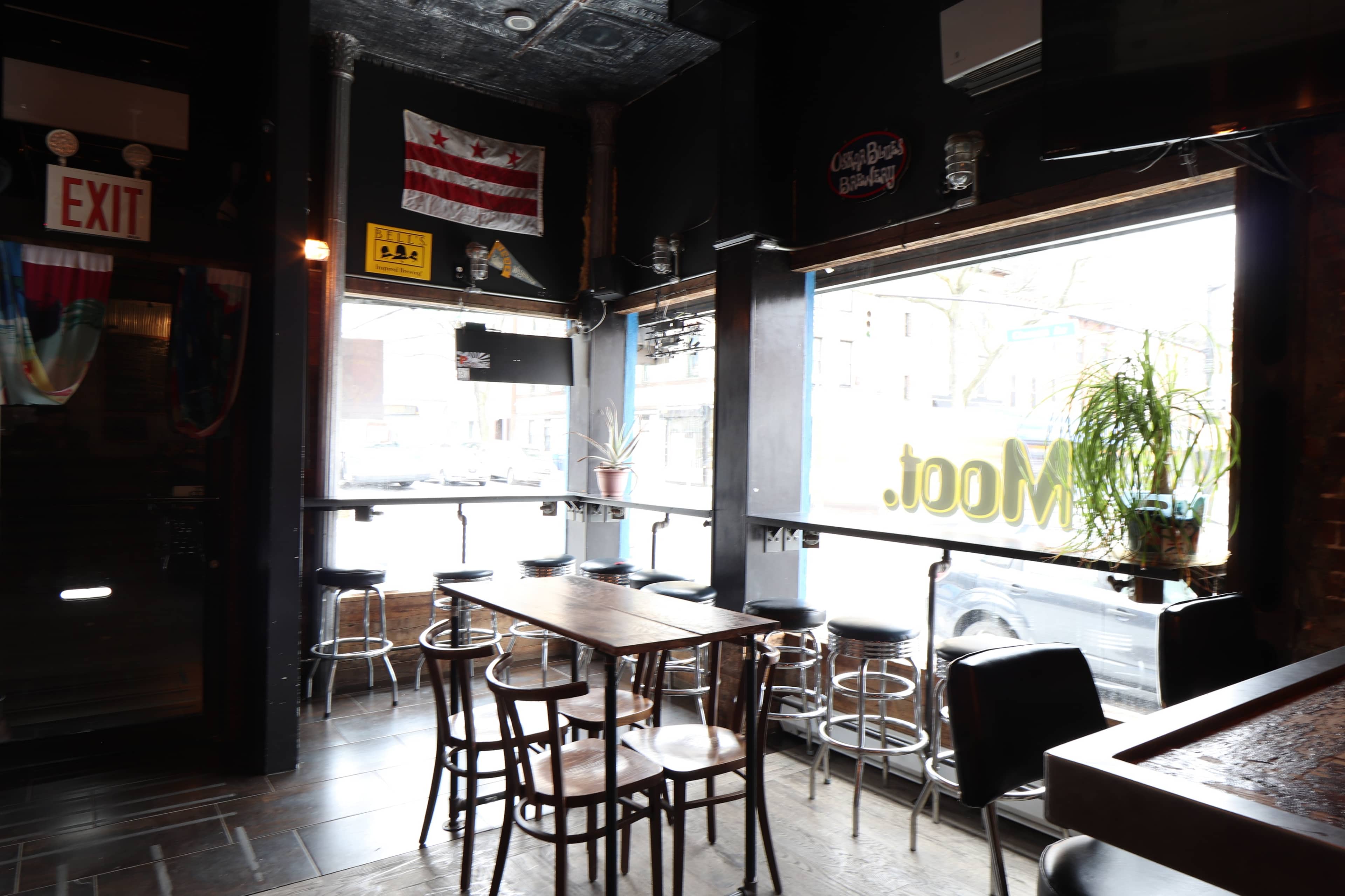 A dimly lit bar interior with a row of bar stools, a wooden table, and large windows displaying a street outside.
