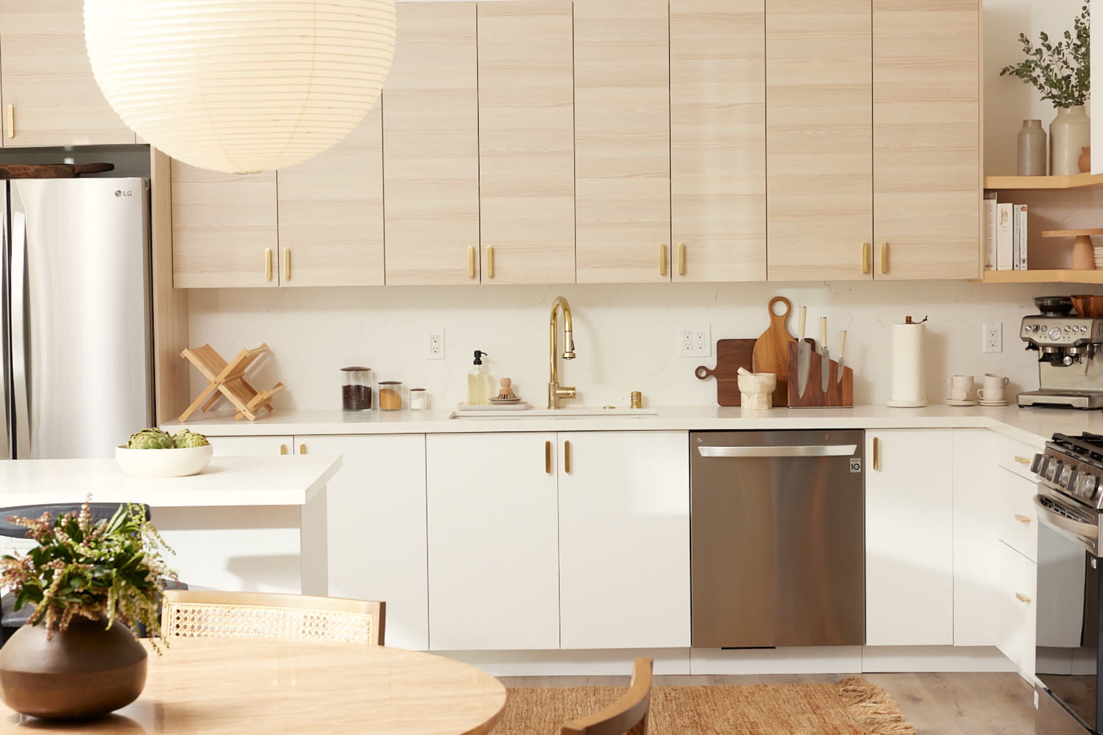 The image shows a modern kitchen with light wood cabinetry, a stainless steel dishwasher, and a round dining table with a centerpiece.