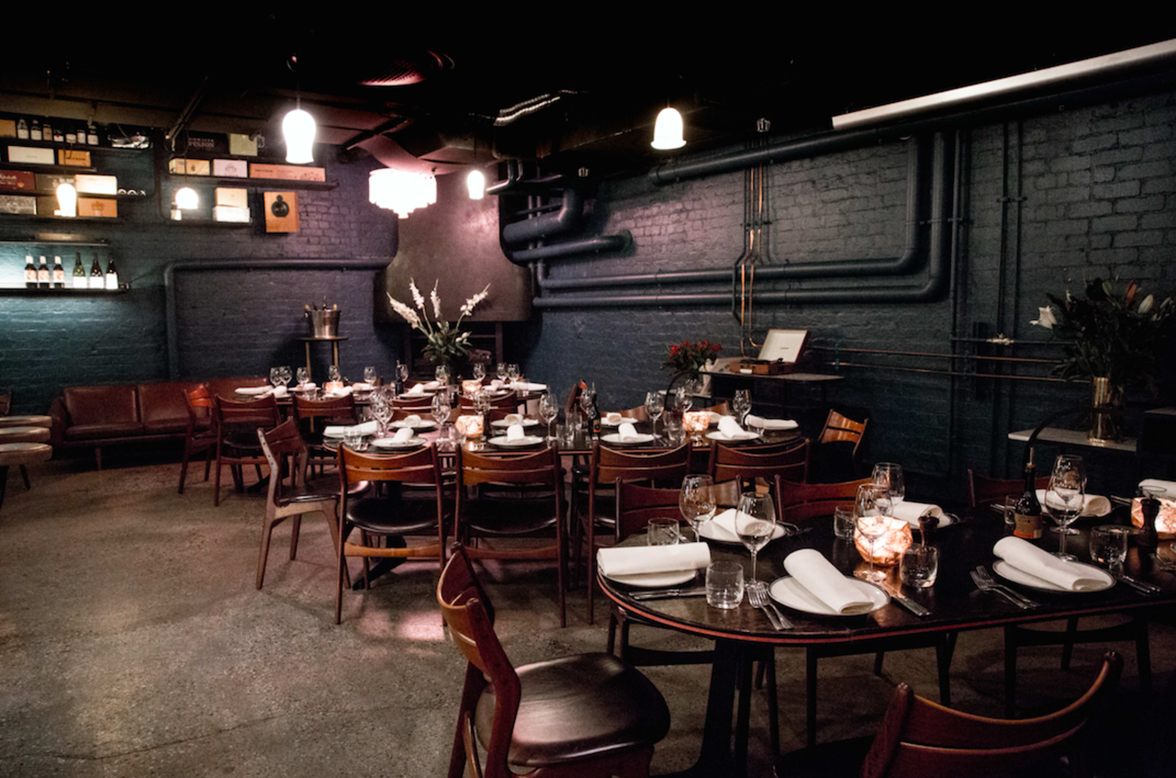A dimly lit restaurant features several wooden tables set with glassware and white napkins against a backdrop of brick walls and industrial pipes.