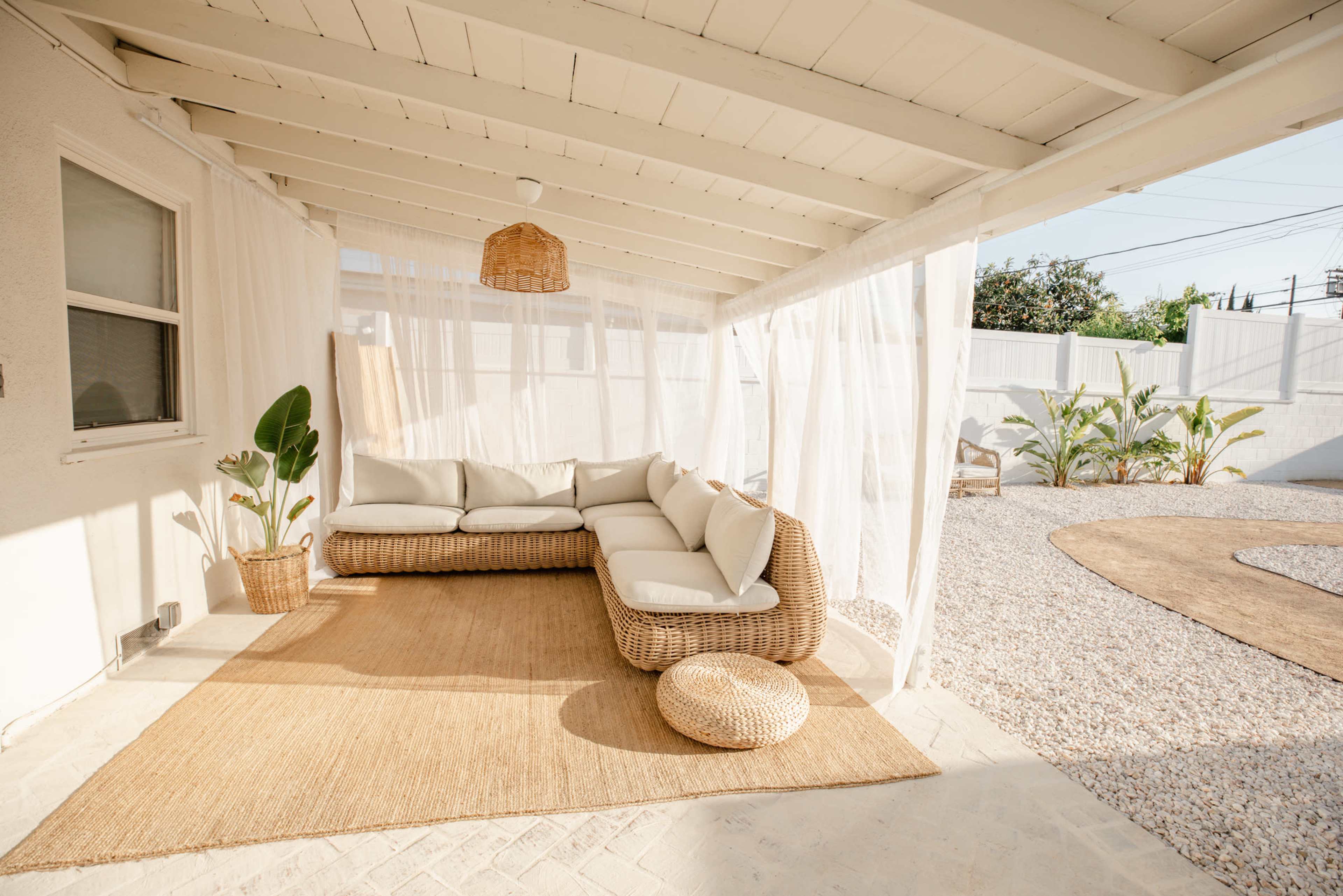 A cozy seating area featuring a sectional couch and a round pouf, set under a covered patio with sheer curtains and a natural fiber rug.